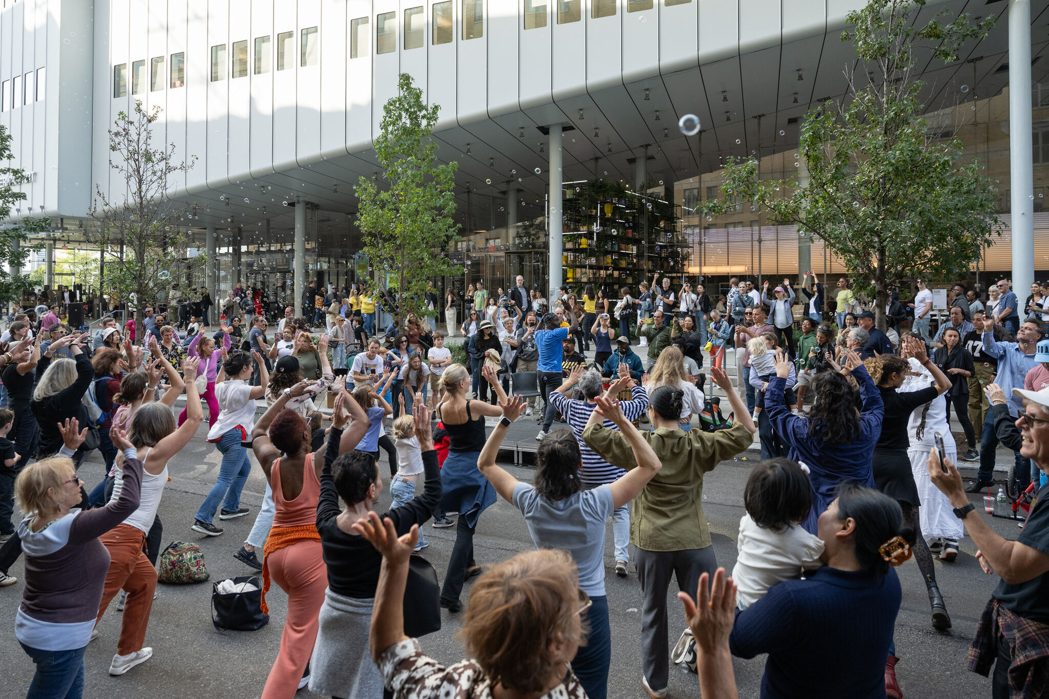 The image shows an intergenerational dance class outdoors at the Whitney Museum during Free Second Sunday. Participants of all ages are engaged in synchronized movements, creating a lively atmosphere. The setting includes greenery, bubbles, and the museum's modern architecture in the background, with a crowd of onlookers and participants enjoying the event.