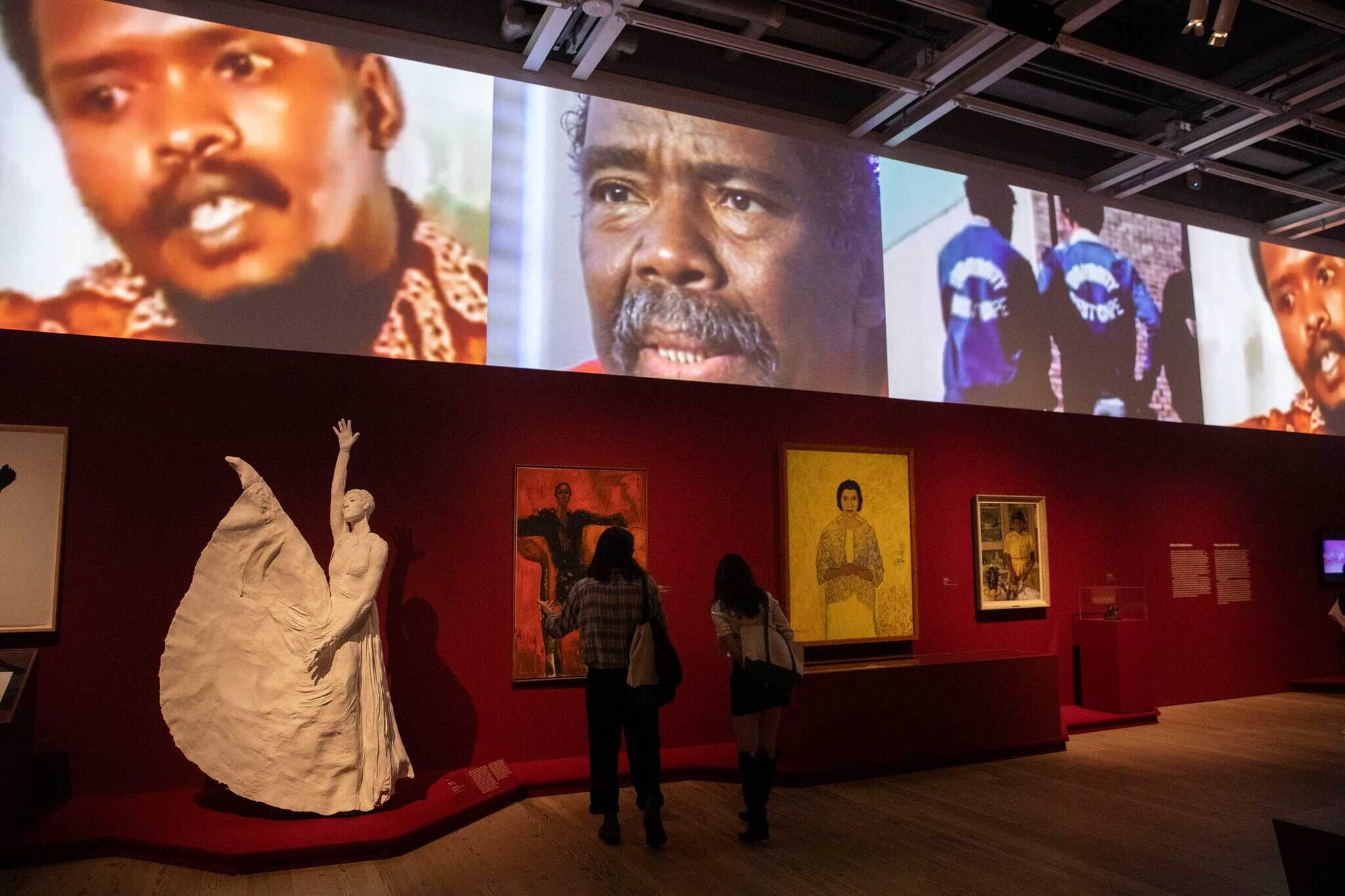 Museum exhibit with a white sculpture, paintings on red walls, and video projections above. Two people observe the artwork.