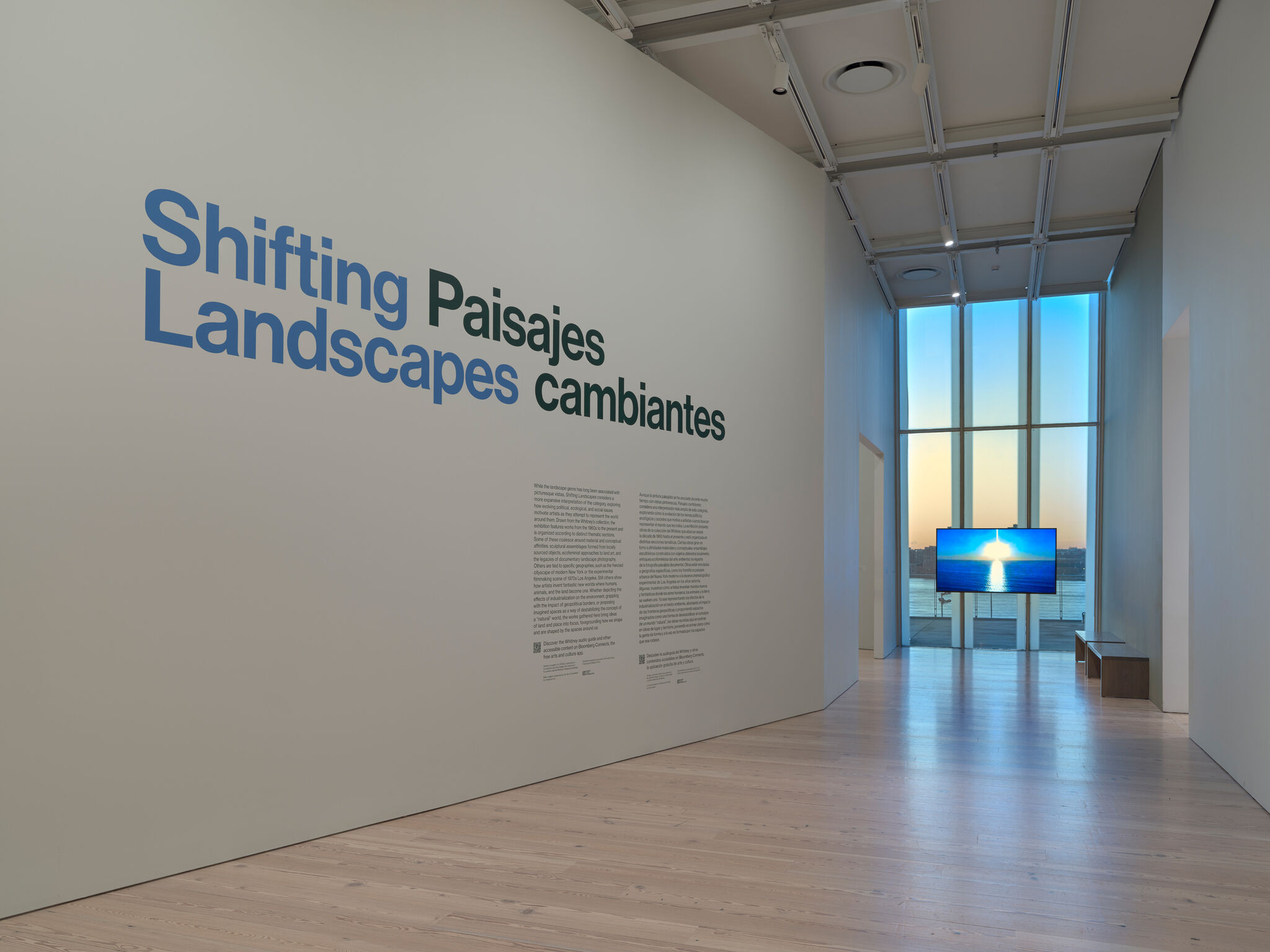 Gallery with "Shifting Landscapes" exhibit text on the wall, a screen displaying a sunset over water, and large windows showing the horizon.