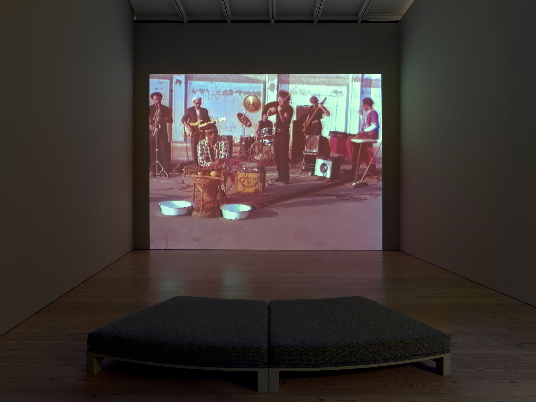 A dark room with a video projection of a band playing instruments. A cushioned bench is placed in front of the screen.