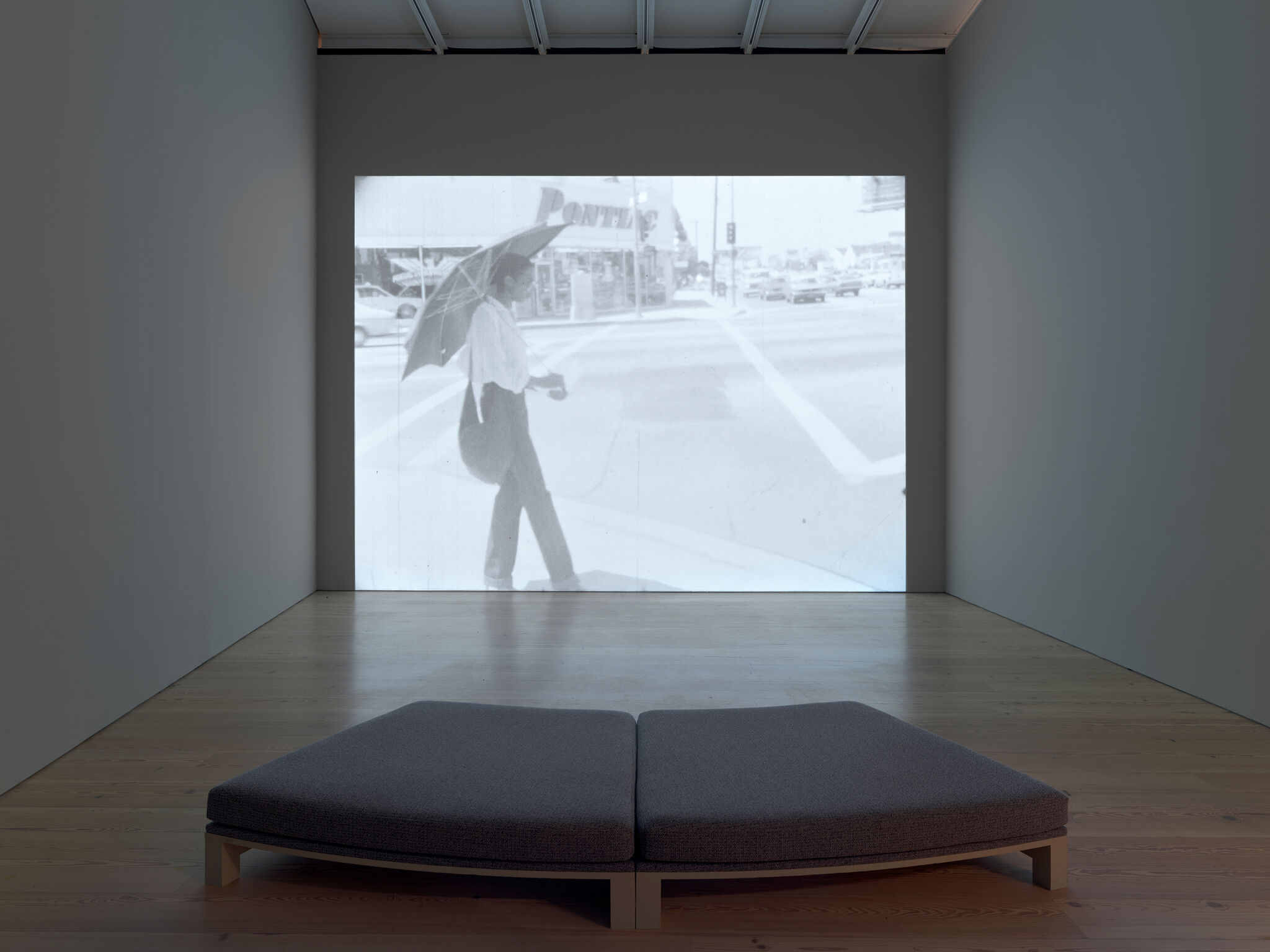 A black-and-white video projection of a person with an umbrella walking on a street corner, displayed in a minimalist gallery space.