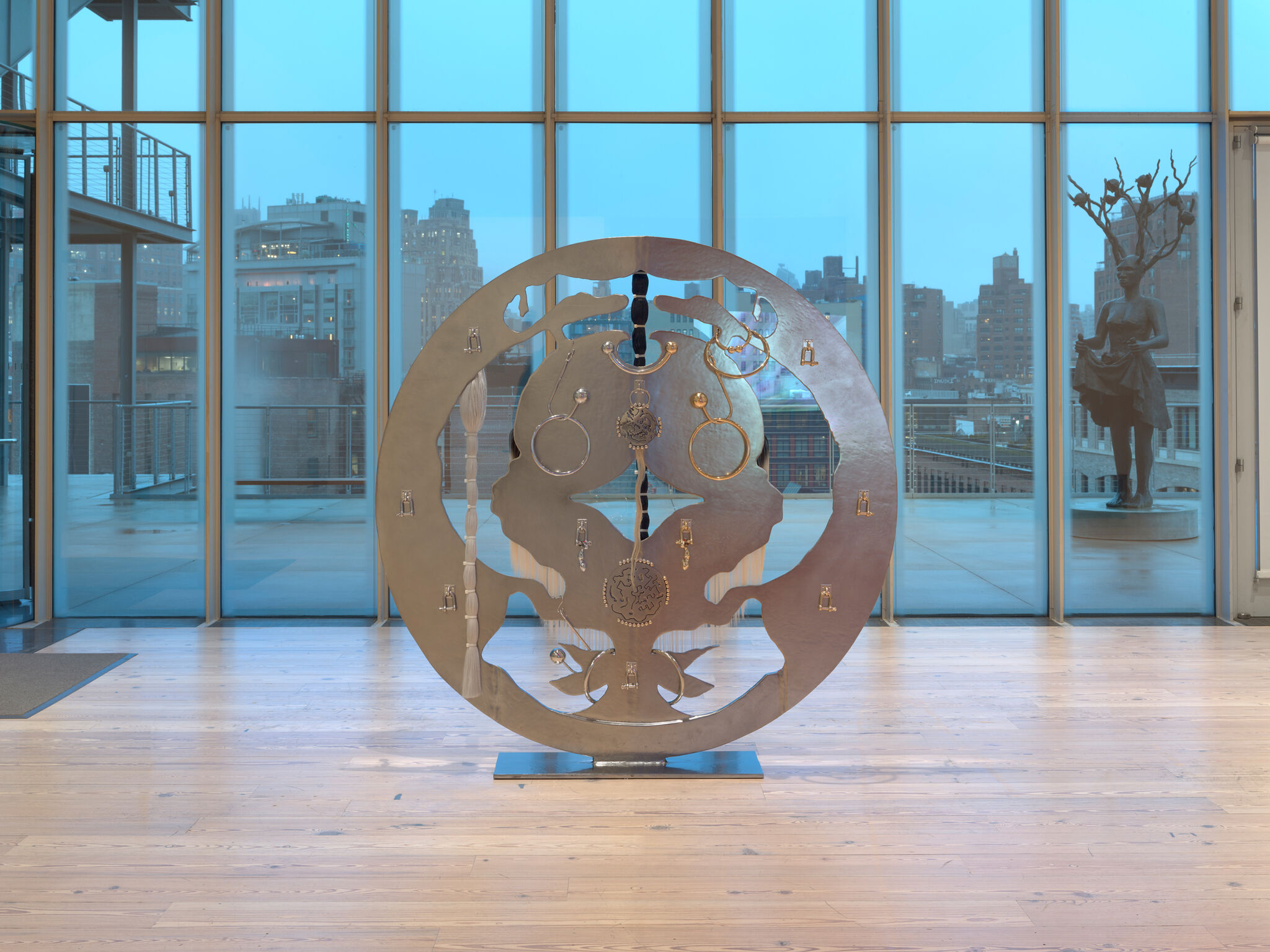 Circular metal sculpture with intricate designs stands in a modern gallery with large windows overlooking a cityscape and a statue outside.