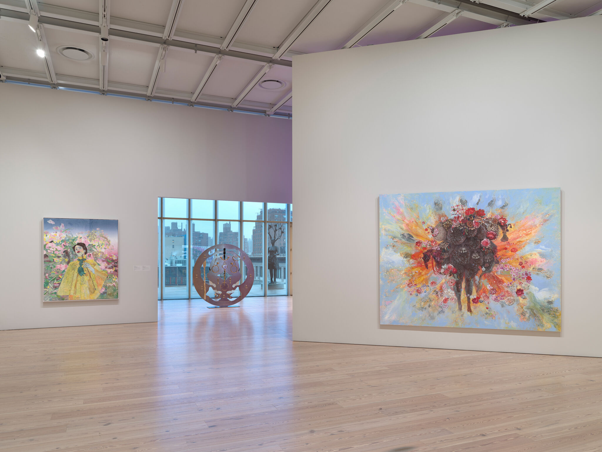 Art gallery with two colorful paintings on white walls, featuring floral and abstract designs. Large windows reveal a cityscape view.