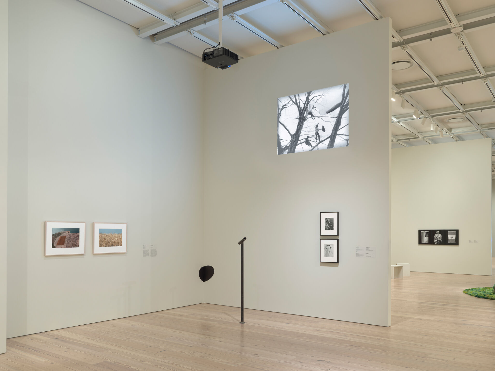 Art gallery with framed photos on white walls, a black-and-white projection above, and a wooden floor. Ceiling lights illuminate the space.