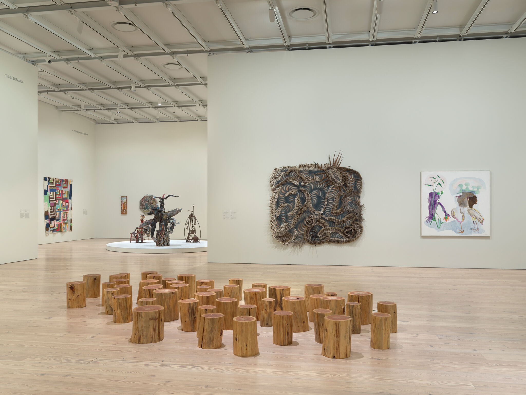 Art gallery with wooden stump installation, abstract wall art, and sculptures on display. Bright, spacious room with light wood flooring.