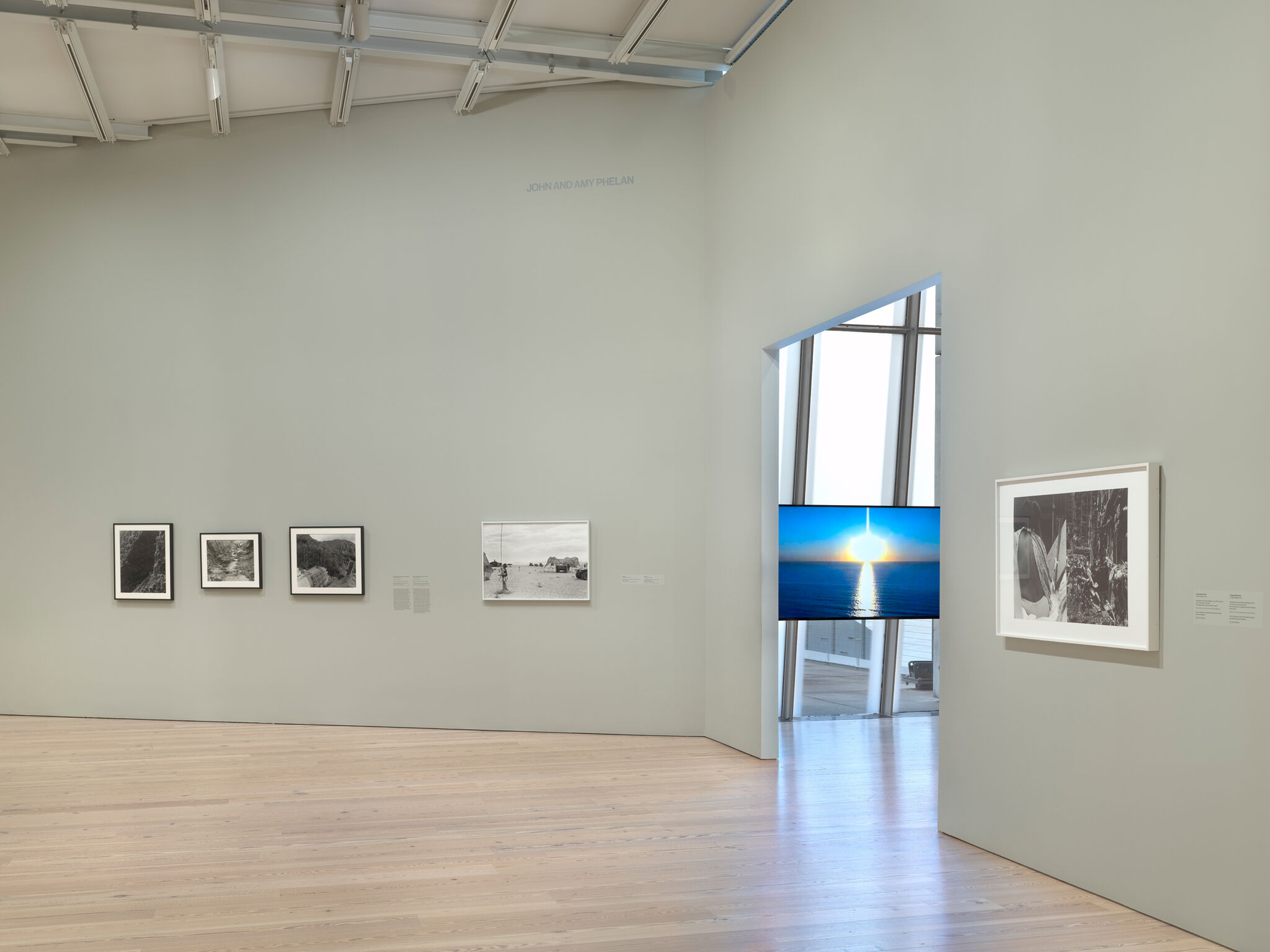 Art gallery with framed black-and-white photos on walls, a digital screen displaying a sunset over water, and a light wood floor.