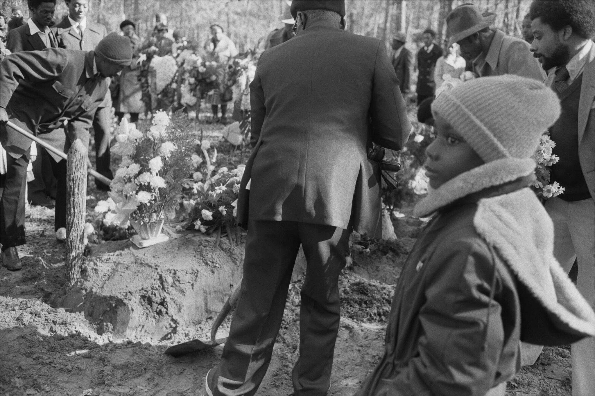 A solemn gathering at a graveside, with people placing flowers and a child in the foreground wearing a winter coat and hat.