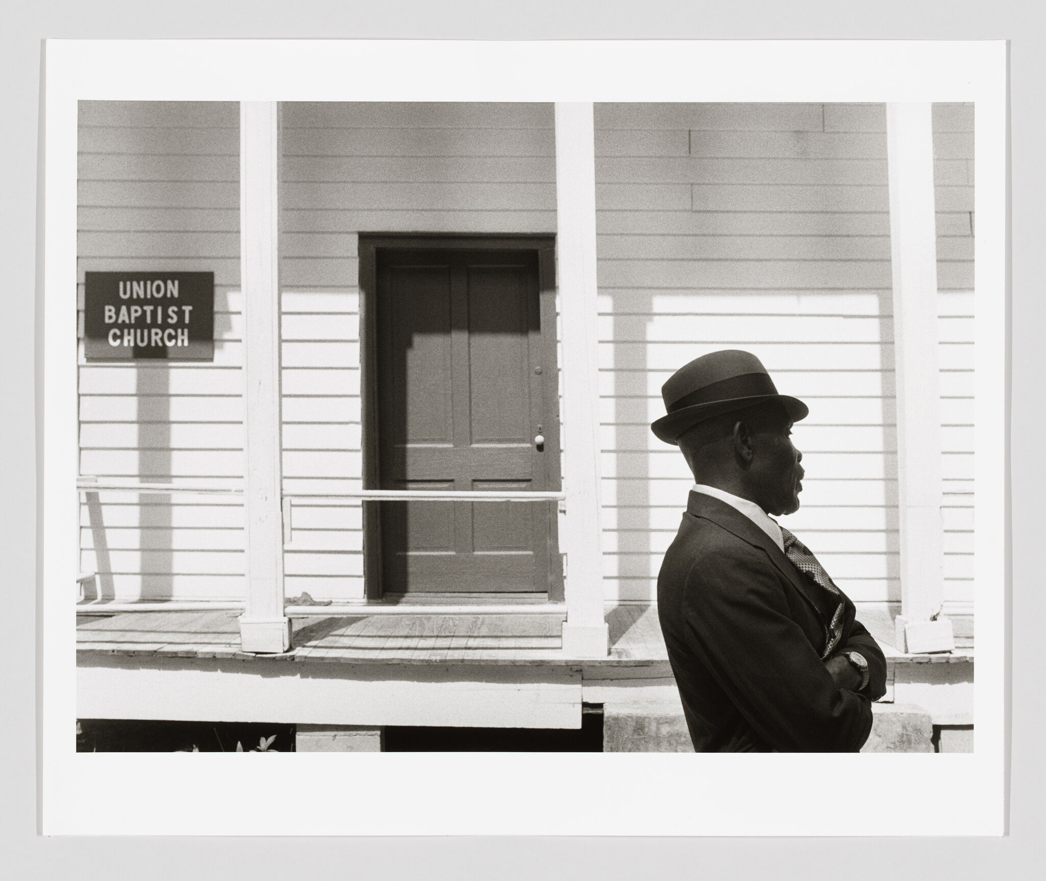 A black-and-white photograph of a man in formal attire, including a suit and hat, standing in profile in front of a white wooden building with a sign that reads "Union Baptist Church." The church has simple architectural details such as columns and a wooden porch.