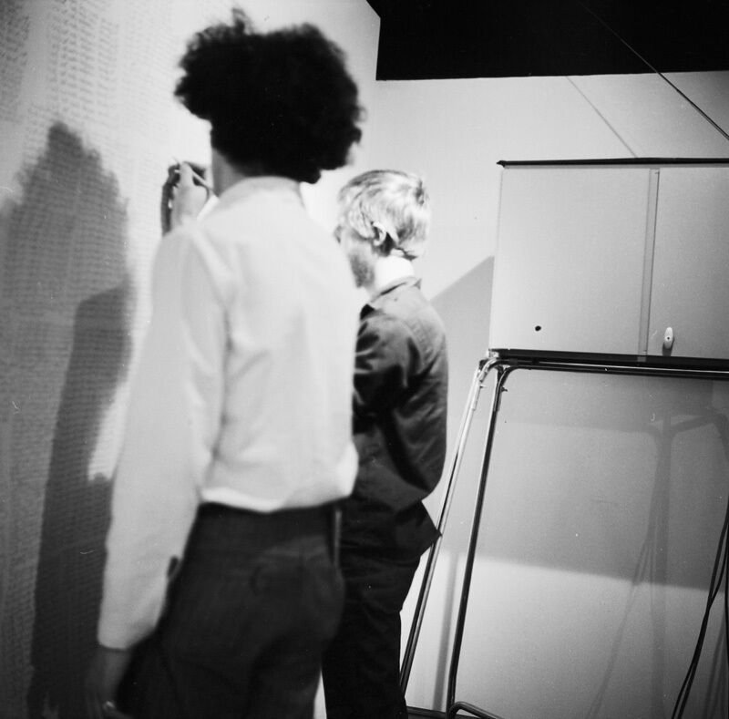 Black and white photo of two people writing on a wall covered in text, with a cabinet and a folding table in the background.