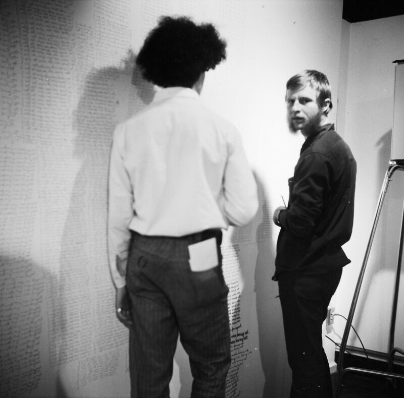 Black and white photo of two people standing in front of a wall covered in handwritten text. One faces away, while the other looks back at the camera.