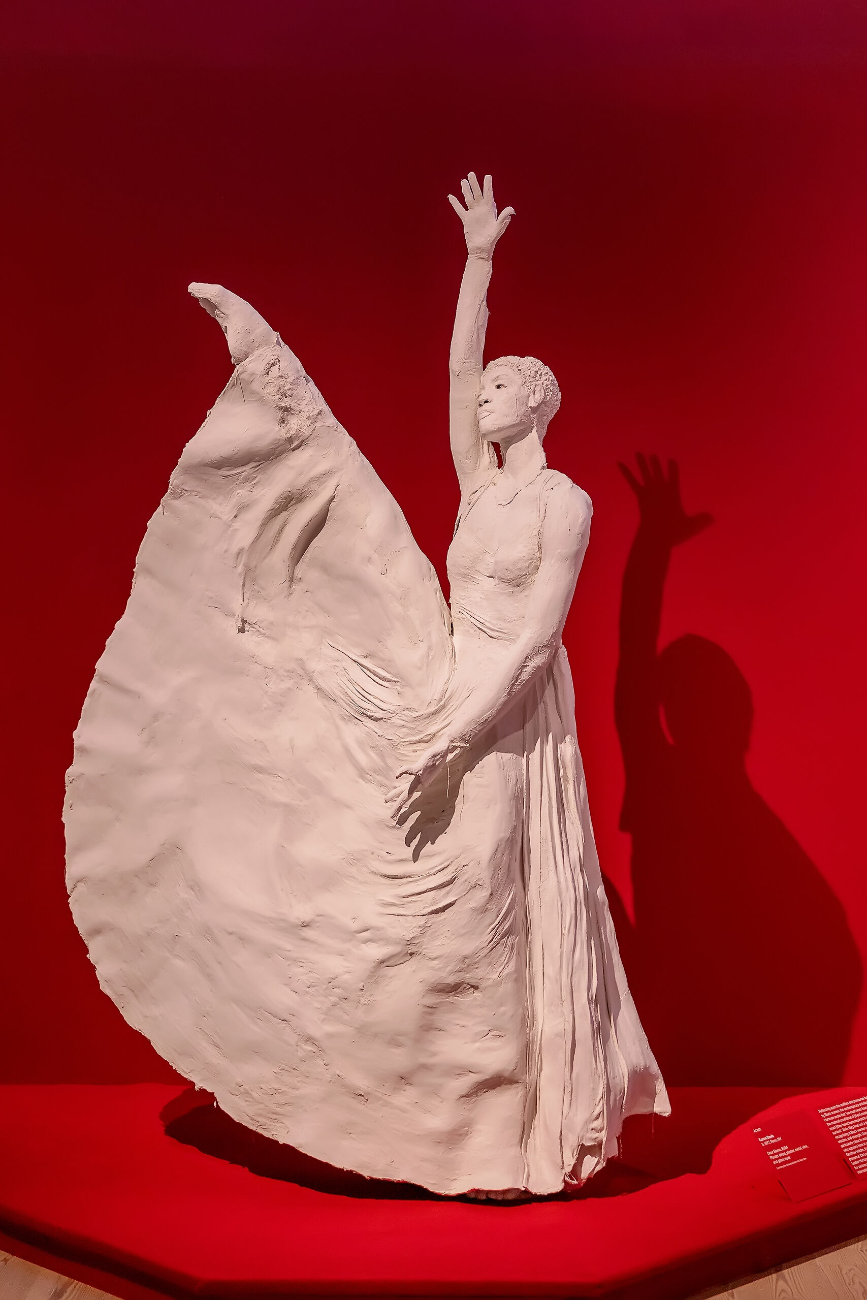 White sculpture of a dancer with an arm raised, set against a vibrant red background. The dancer's shadow is visible on the wall.