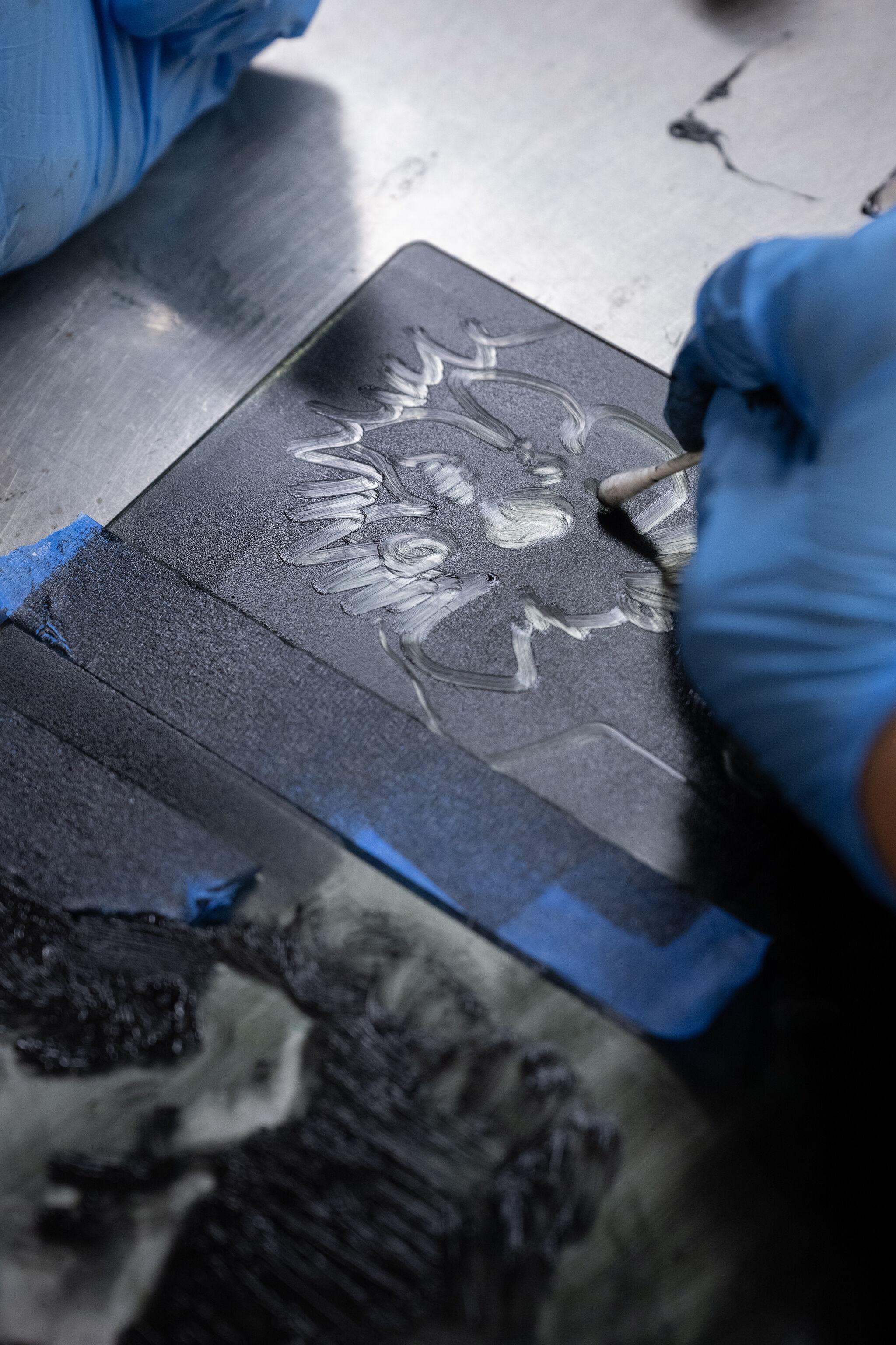 A close-up of a gloved hand using a cotton swab to create intricate designs on a dark, textured surface. The process appears to be part of a printmaking or engraving activity, with the surface taped down for stability. The artistic details and hands-on technique suggest a workshop or educational setting focused on creative expression.