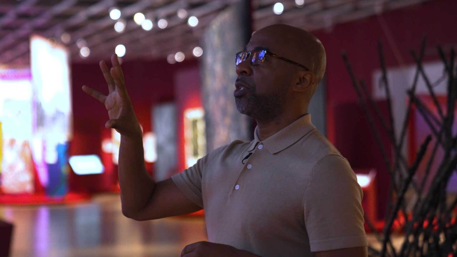 A person in a beige shirt gestures while speaking in a dimly lit room with colorful displays and abstract decor in the background.