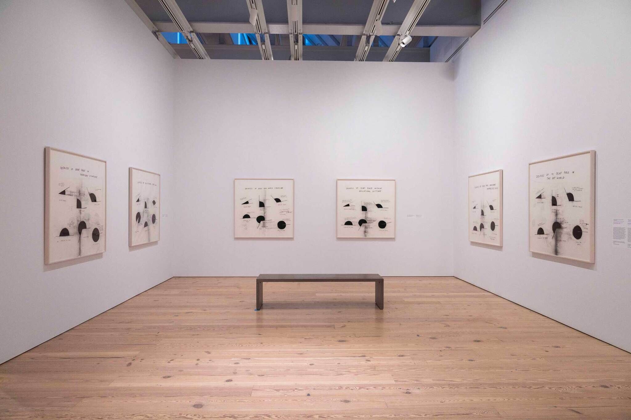 Art gallery room with six abstract framed artworks on white walls, centered bench on wooden floor, and overhead lighting.