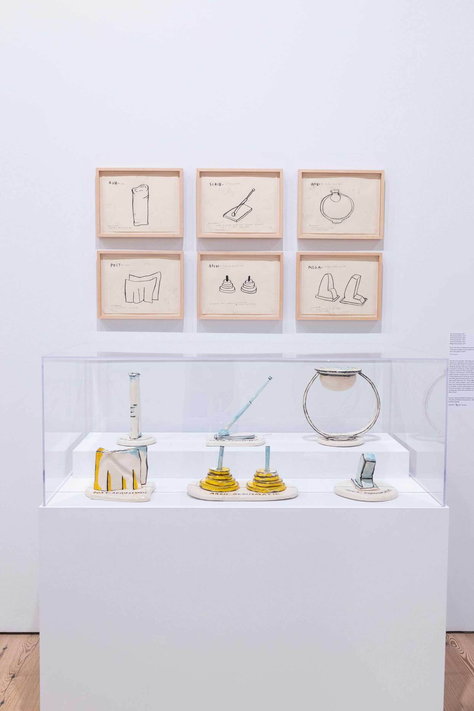 Art exhibit with six framed sketches above a display case containing abstract ceramic sculptures on a white pedestal.