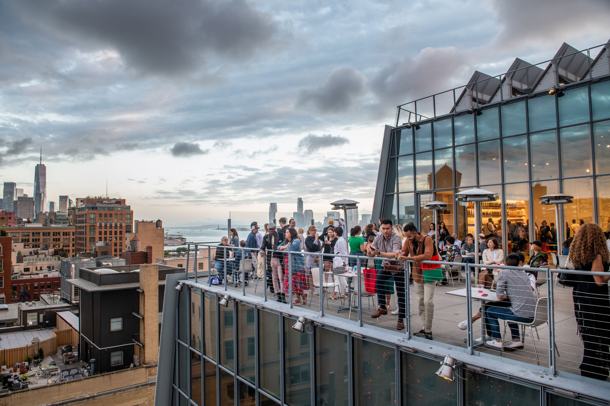 People socializing on a rooftop terrace with city skyline and waterfront views in the background, under a cloudy sky.