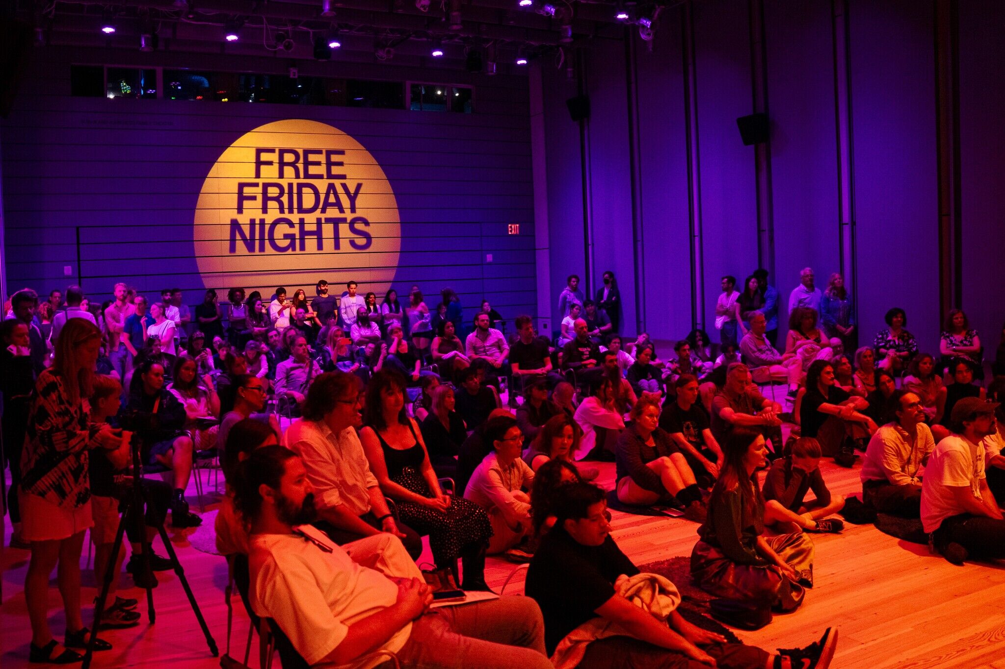 A crowd sits and stands in a dimly lit room with purple lighting. A large yellow circle on the wall reads "Free Friday Nights."