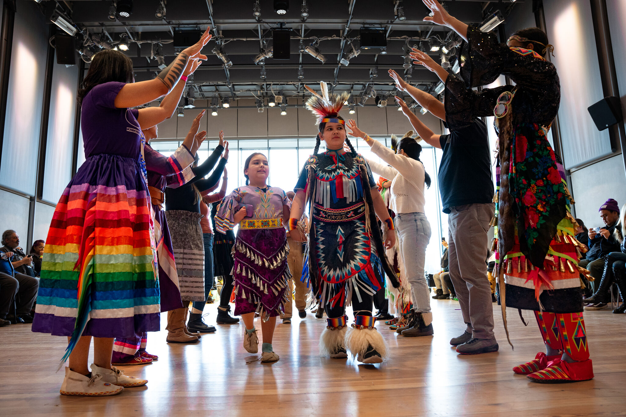 Two children dressed in regalia walking through a path of adults with their arms raised who are also dressed in regalia