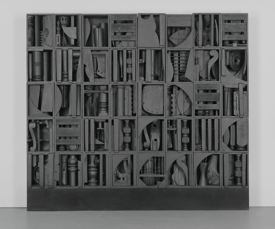 A black wooden assemblage sculpture composed of various geometric and abstract shapes arranged in a grid pattern against a white wall.