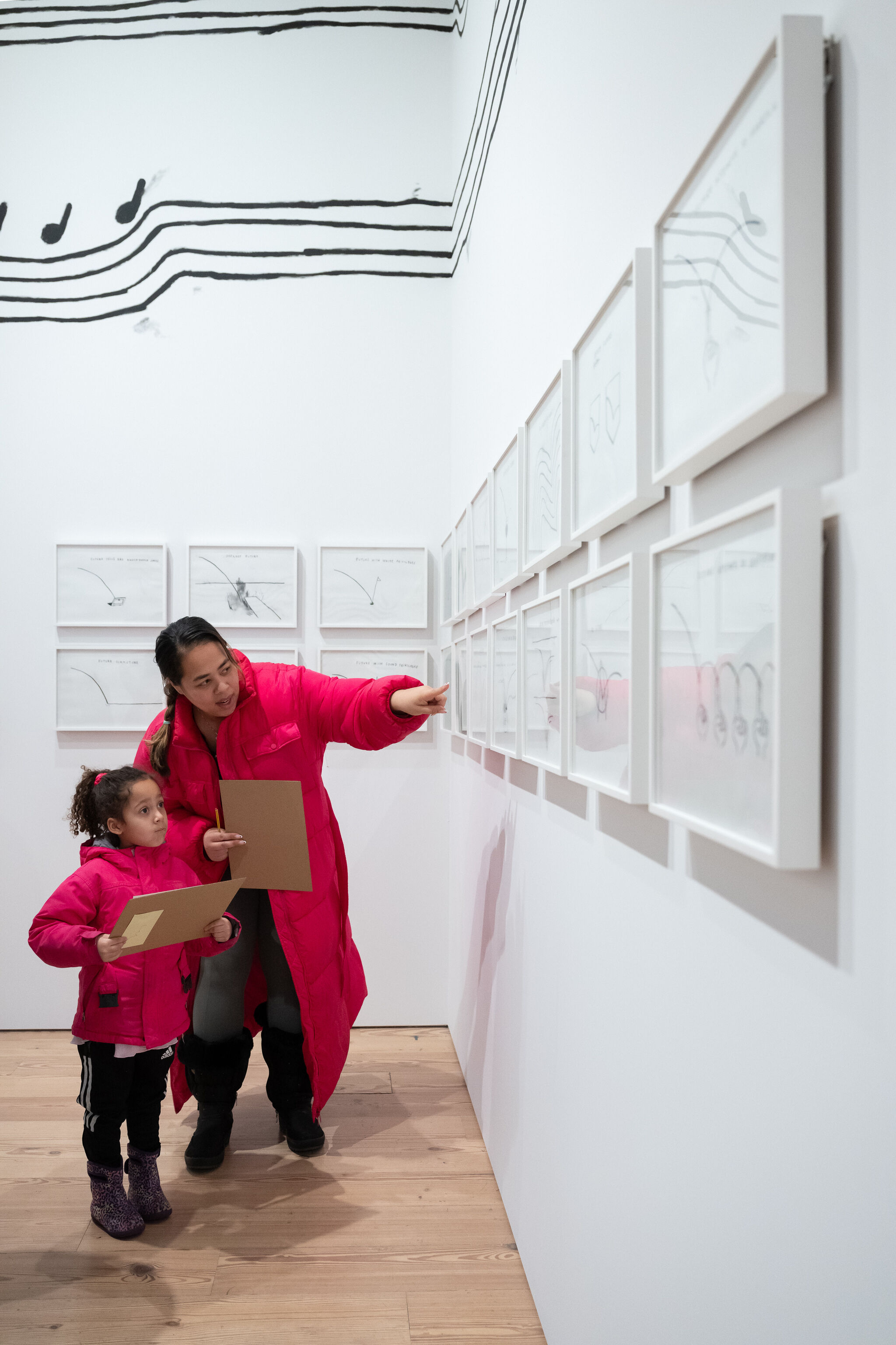 An adult and a child, both wearing matching bright pink winter coats, explore All Day, All Night, an exhibition featuring the artwork of Christine Sun Kim. The adult gestures toward one of the framed drawings on the white gallery wall, engaging the child in conversation about the piece. The child, holding a clipboard, observes the artwork with focus. The exhibition space showcases minimalist black-and-white line drawings, some incorporating musical notation, with a mural of musical notes and curved lines extending onto the upper part of the wall. The interaction highlights curiosity, learning, and engagement with the artist’s work.