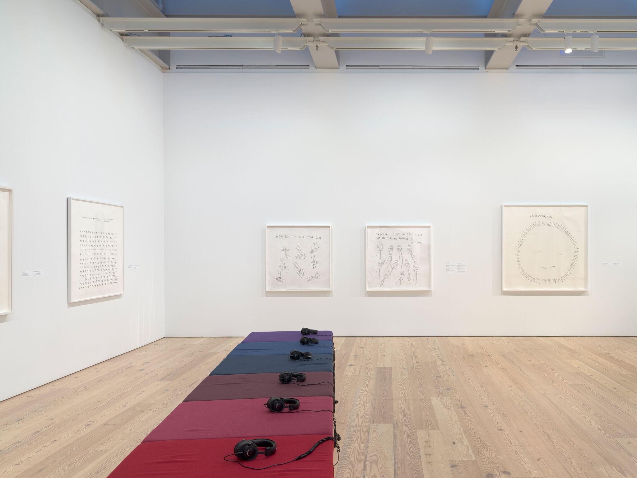 Art gallery with framed sketches on white walls. A row of colorful cushions with headphones is placed on the wooden floor.