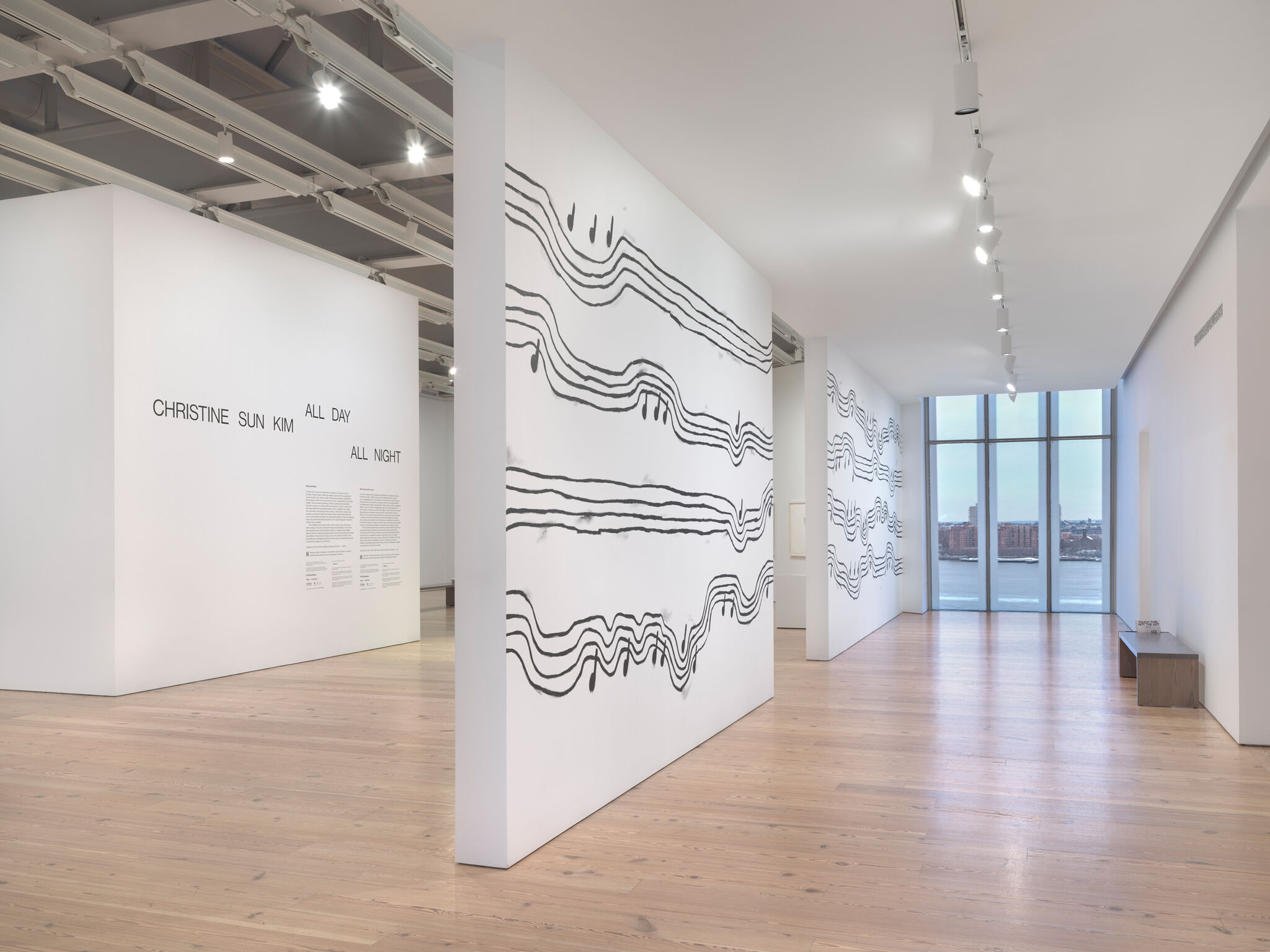 Art gallery with white walls featuring black wavy lines and musical notes. Large windows overlook a river. Text reads "Christine Sun Kim."