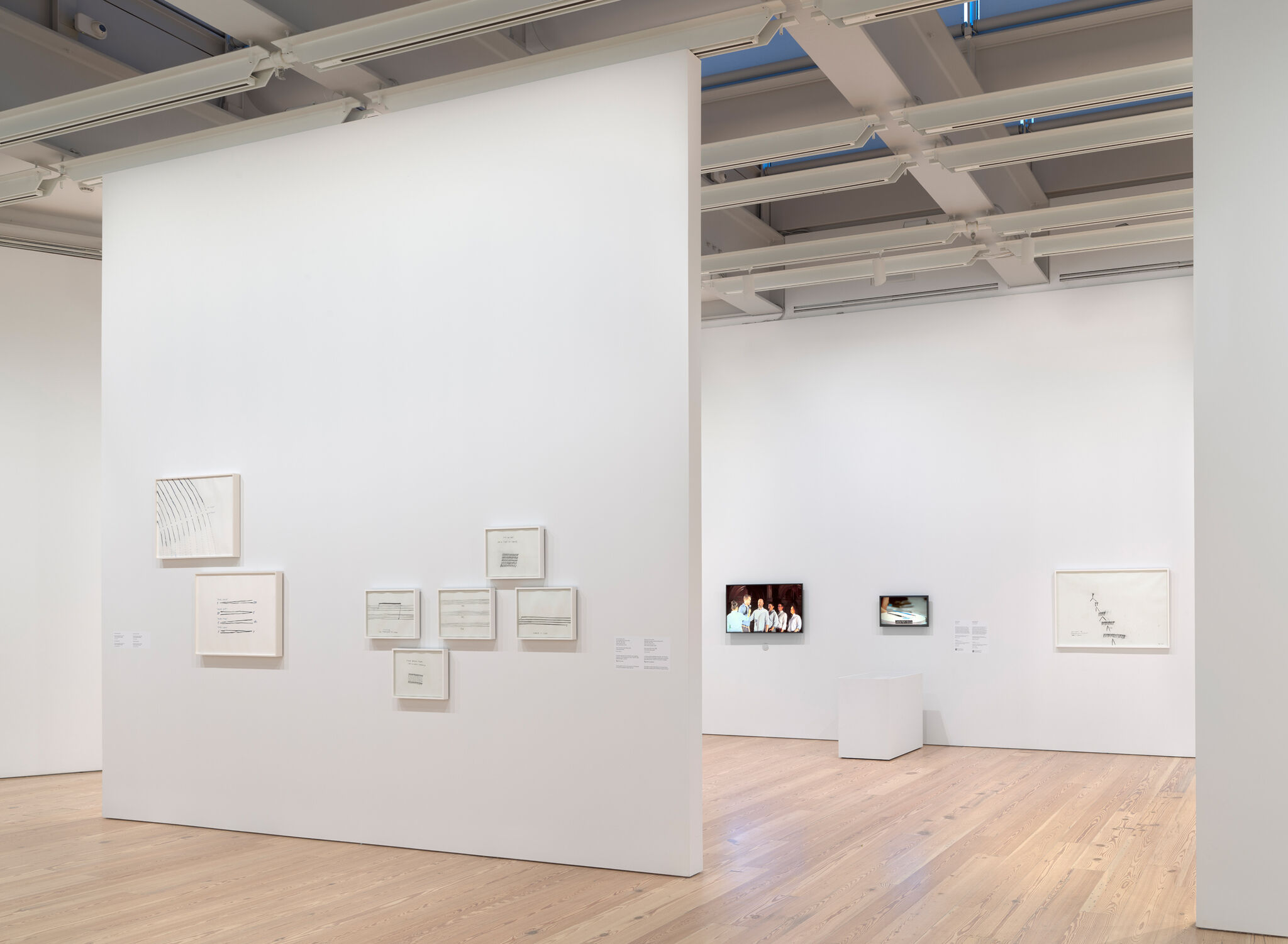 Art gallery with framed drawings on white walls, wooden floor, and two video screens displaying images.