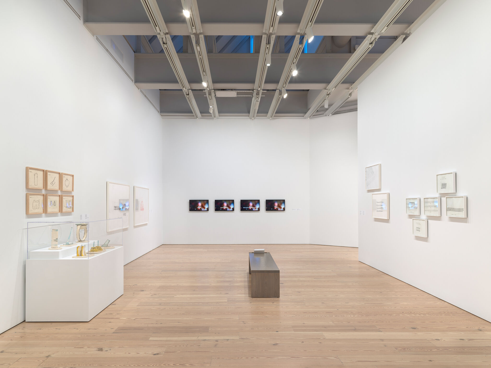 Modern art gallery with framed artworks on white walls, a display case with objects, and a wooden bench on a light wood floor.