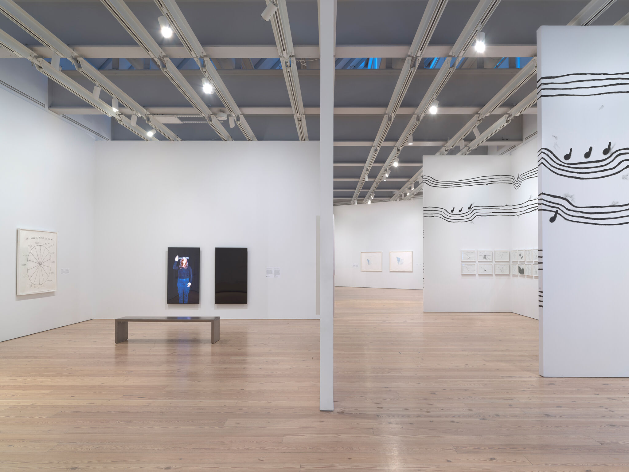 Spacious art gallery with wooden floors, featuring abstract paintings and a bench. Walls adorned with musical note patterns.