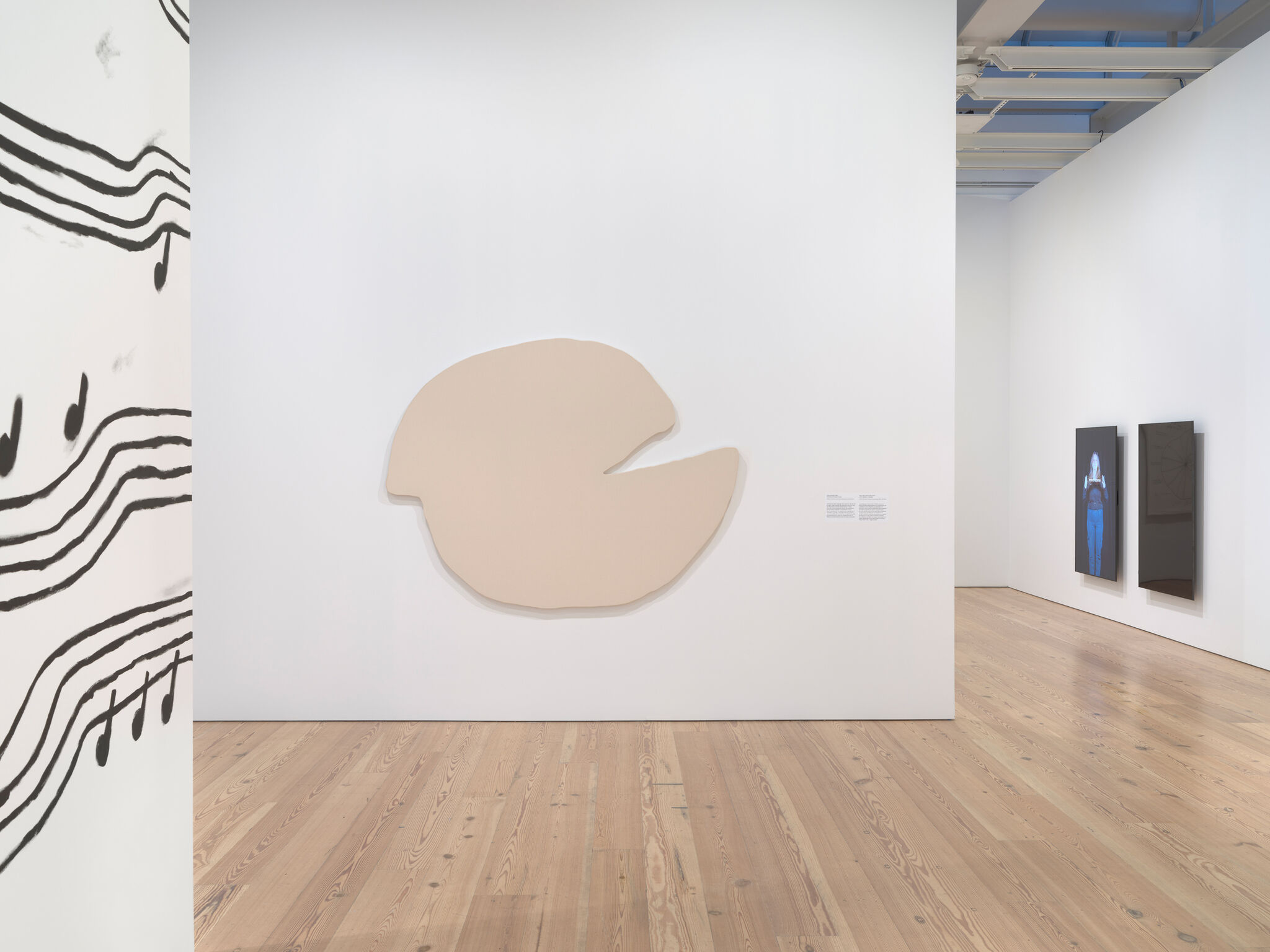 Art gallery with abstract beige shape on wall, musical notes on left, and two digital screens on right. Light wood flooring.