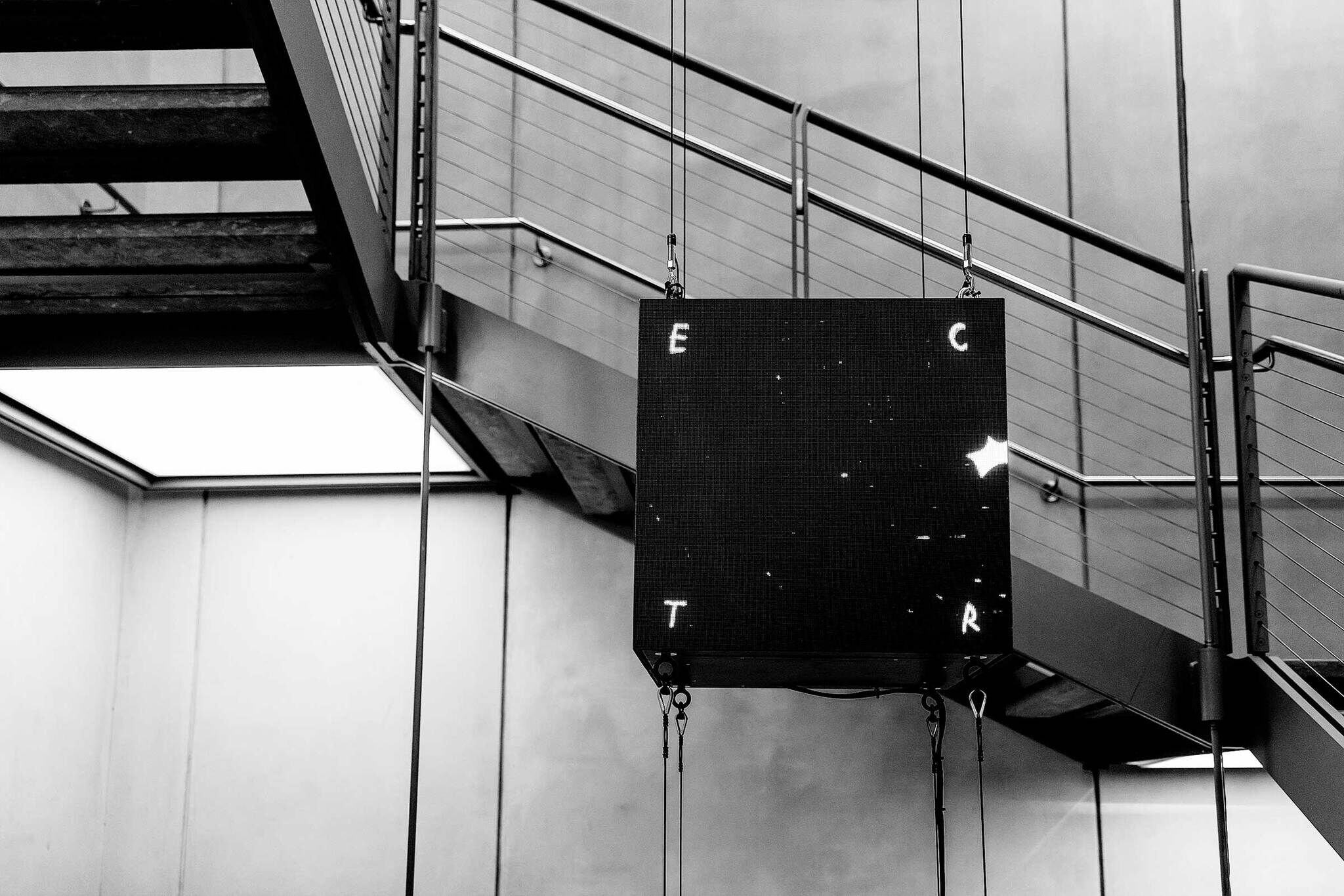 Black and white image of a suspended digital screen displaying scattered letters "E," "C," "T," "R" under a modern staircase.