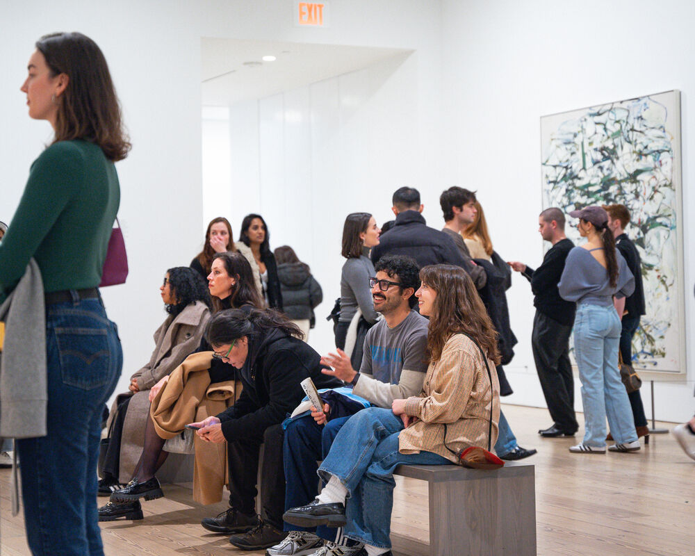 People in a gallery, some seated and others standing, engage with art and each other. A large abstract painting is visible on the wall.