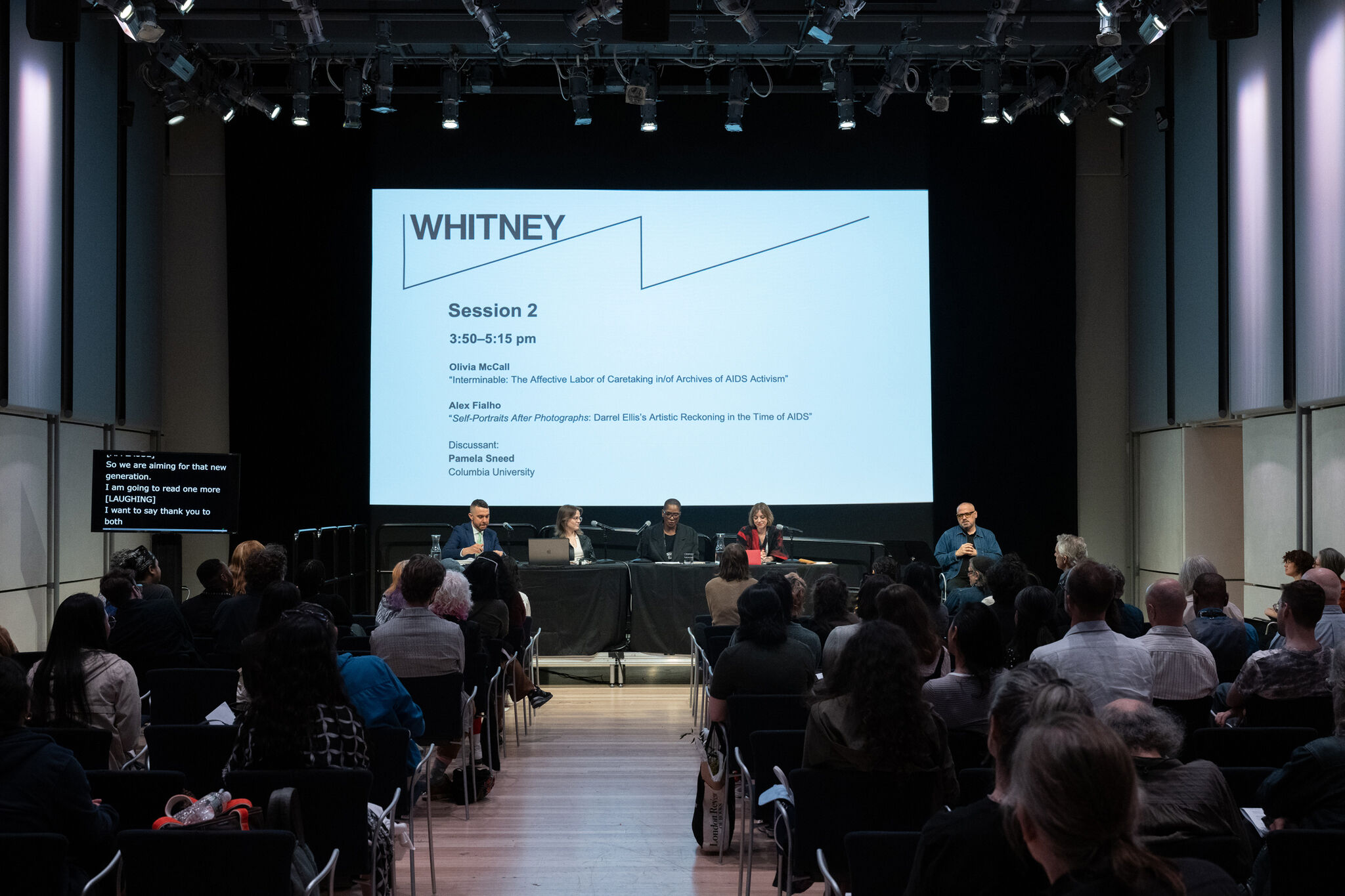 A panel discussion at the Whitney Museum of American Art features four speakers seated at a long table on a stage, with a large screen behind them displaying the event title 'Session 2' and presentation topics related to AIDS activism and artistic reckoning. The screen also lists the names Olivia McCall, Alex Fialho, and Pamela Sneed. A man in a blue shirt stands to the right, speaking into a microphone. The audience, consisting of a diverse group of attendees, is seated in rows facing the stage. The room is well-lit with overhead spotlights illuminating the stage.
