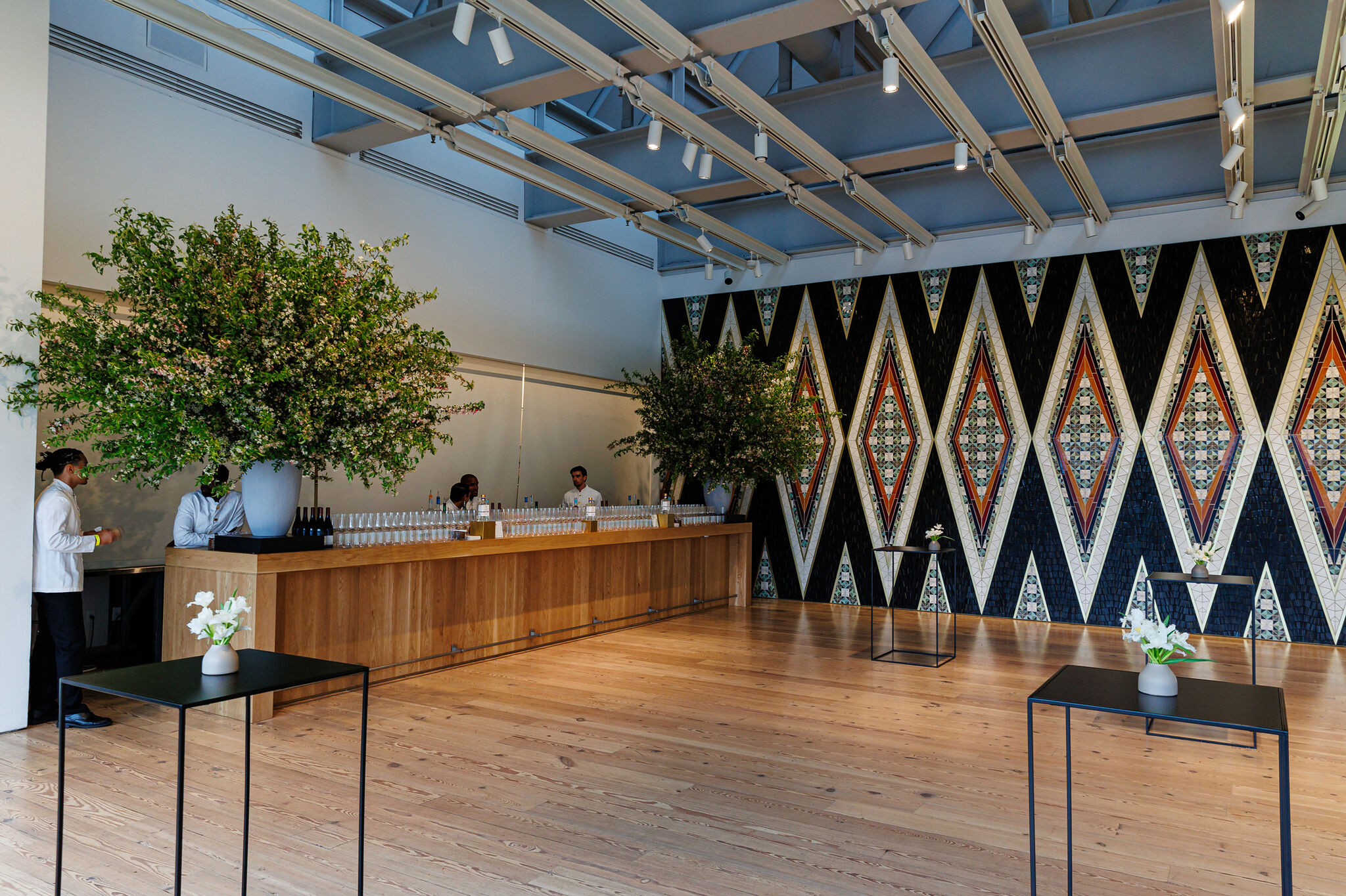 Modern room with geometric wall art, large floral arrangements, wooden bar, and small tables with vases on a wooden floor.