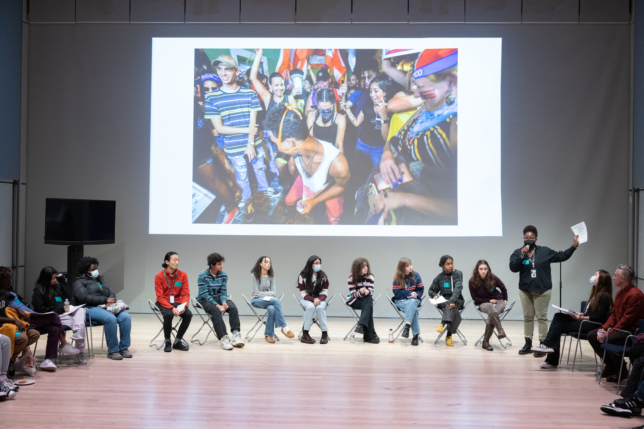 A group of people sit in a semi-circle on stage, with one person standing and speaking. A large photo of a lively crowd is projected behind them.