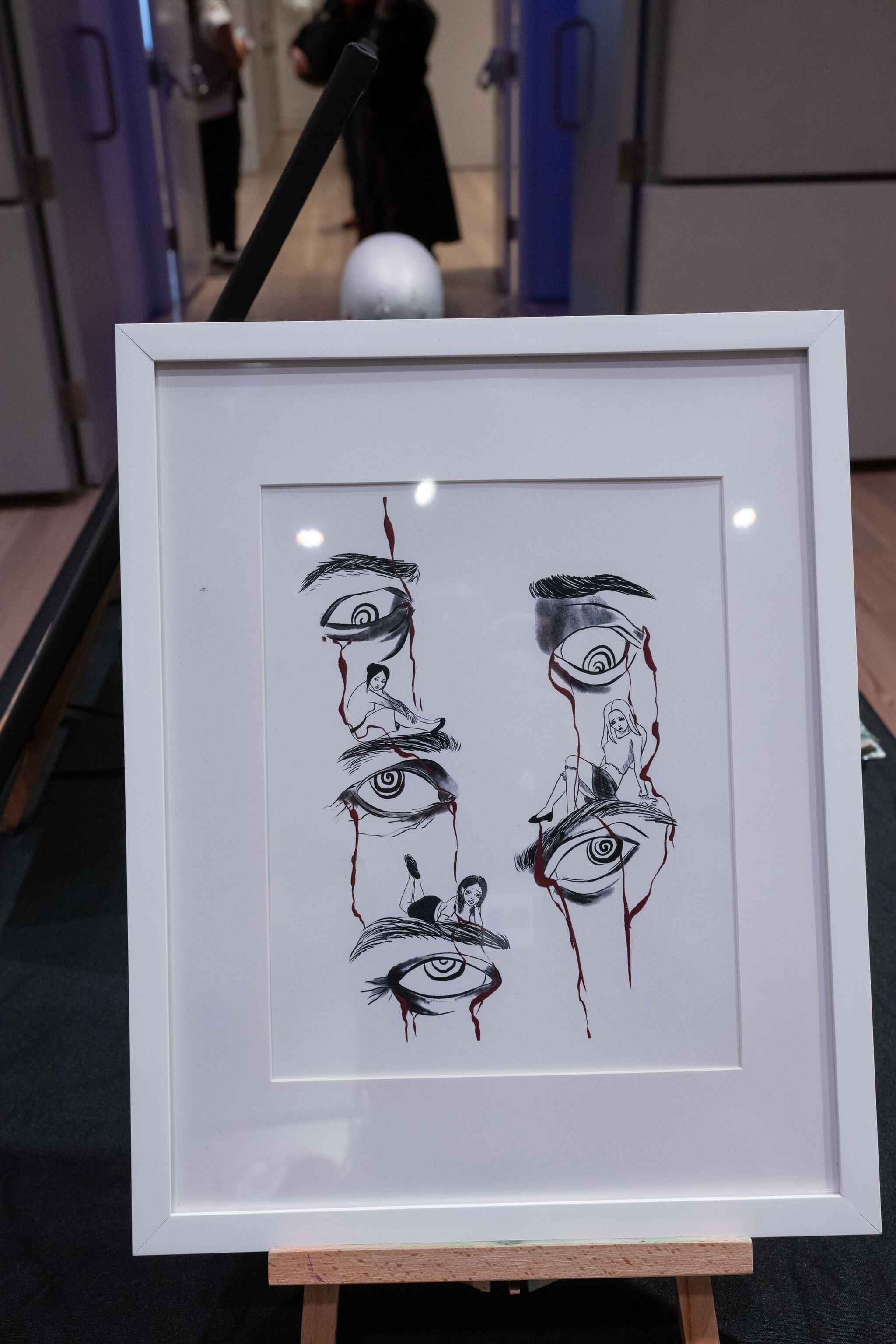 Framed artwork featuring eyes with red lines and small figures sitting on them, displayed on an easel in a room.