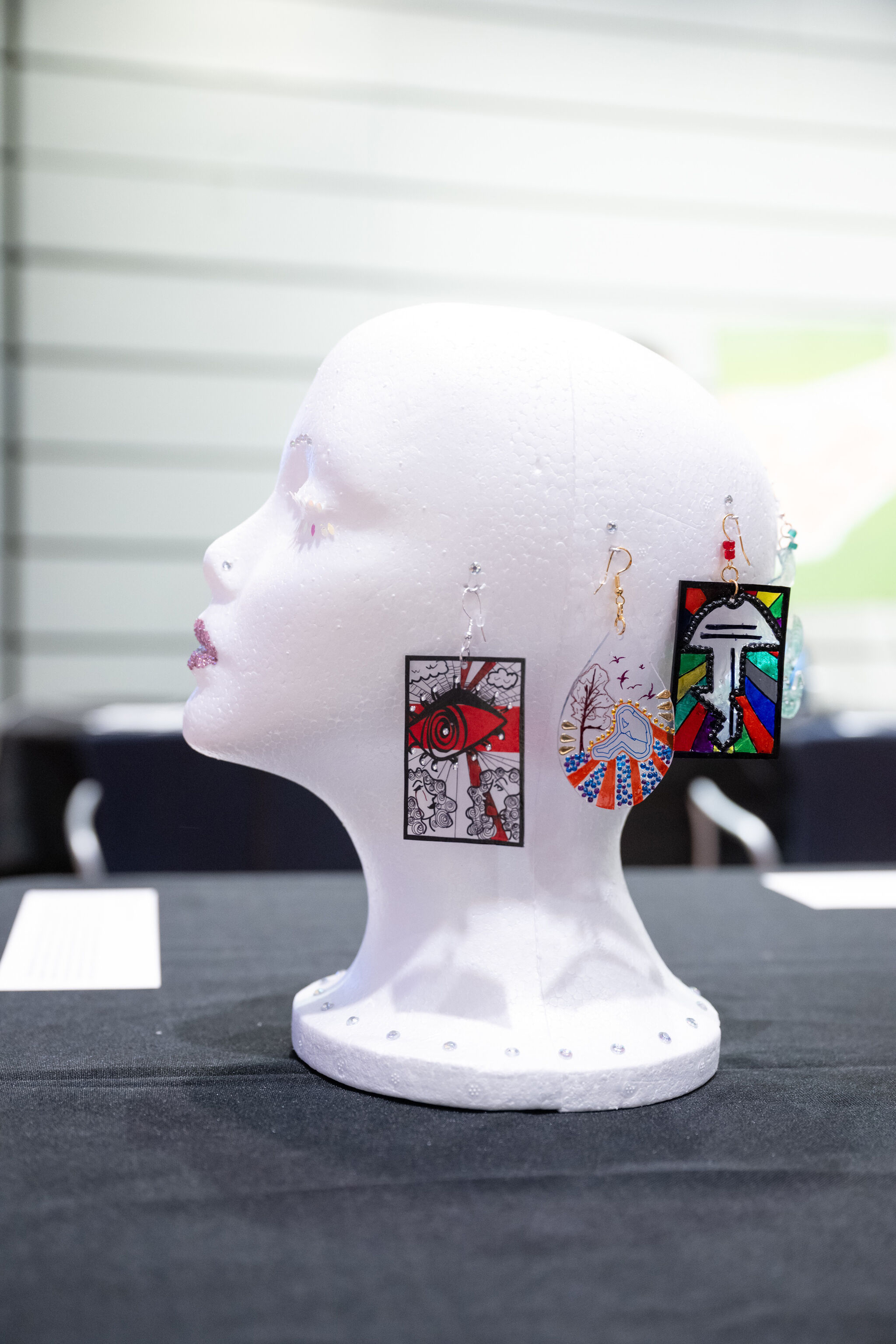 White mannequin head displaying colorful, artistic earrings on a black table, with a blurred background.