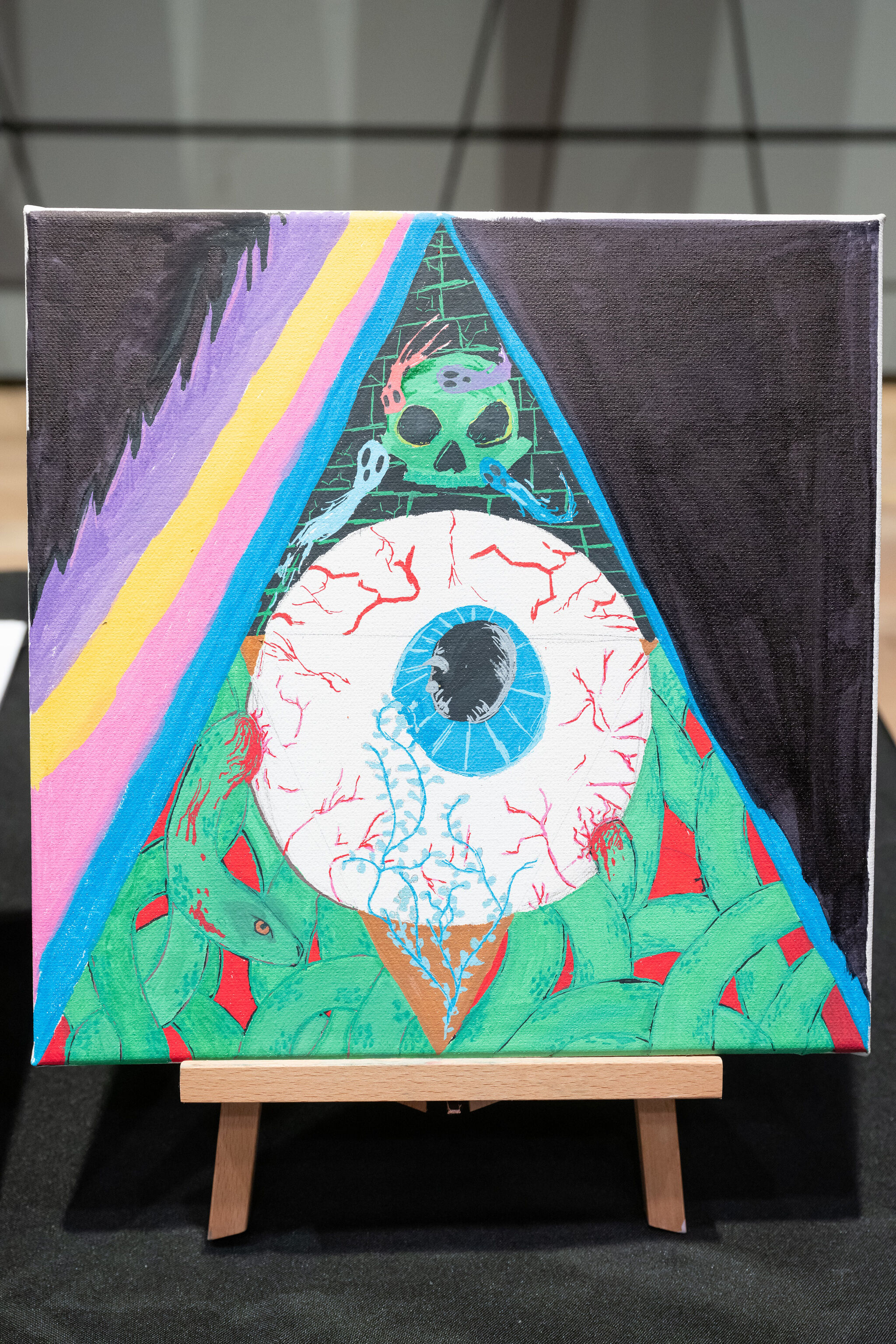 Colorful painting of a large eye within a triangle, surrounded by snakes, a skull, and abstract patterns on a canvas stand.