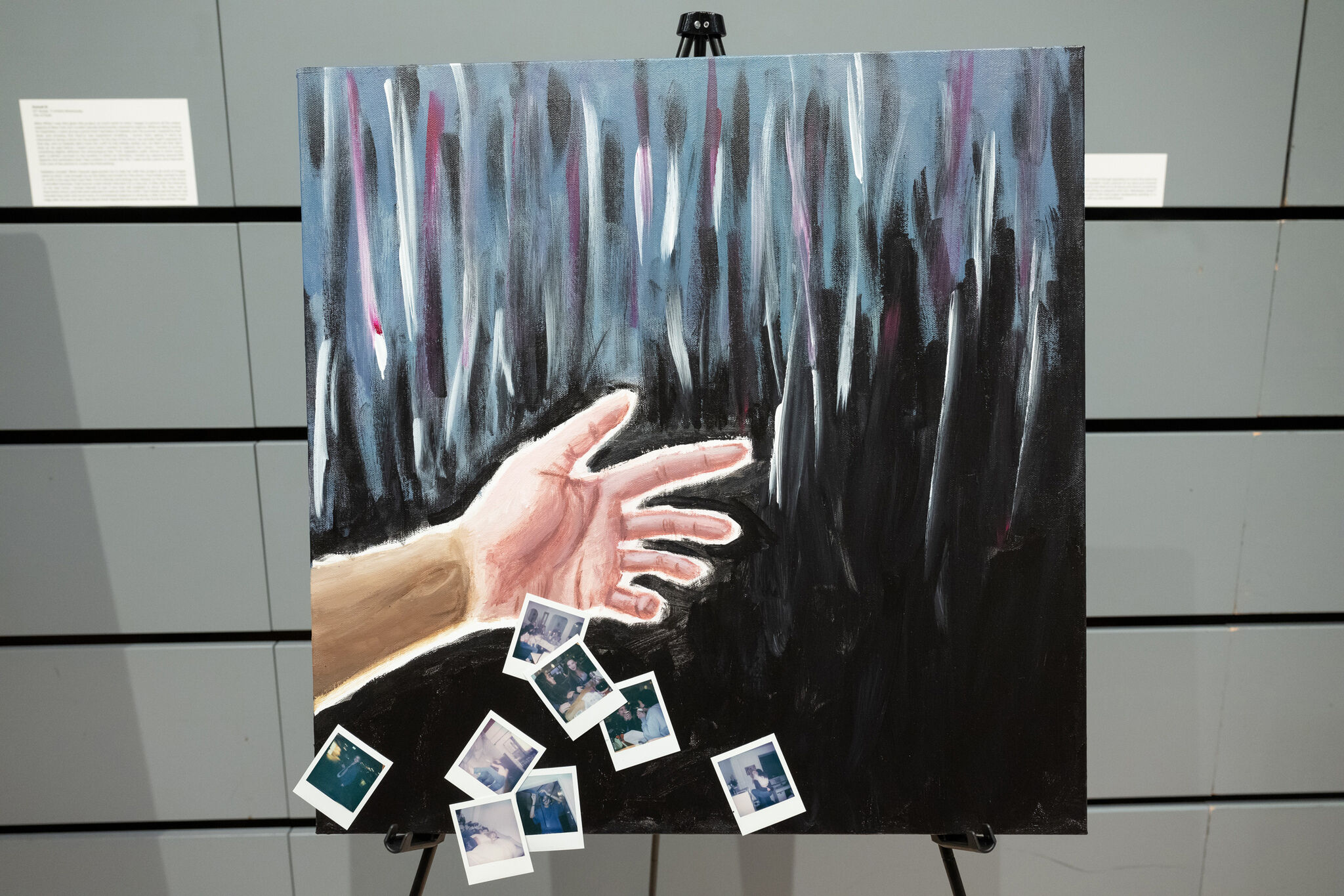 Painting of an outstretched hand against a dark, abstract background with scattered Polaroid photos at the bottom.