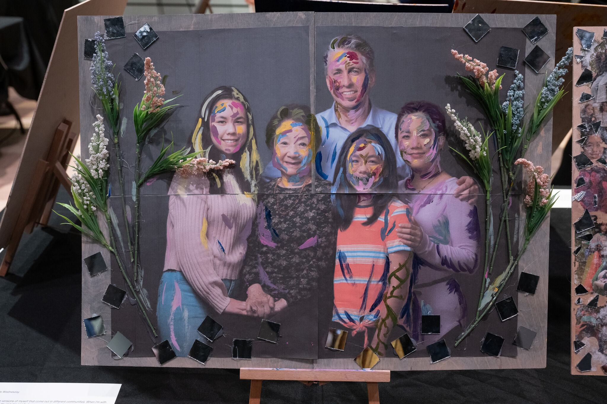 Family portrait with colorful paint strokes on faces, surrounded by flowers and mirrored tiles, displayed on an easel.