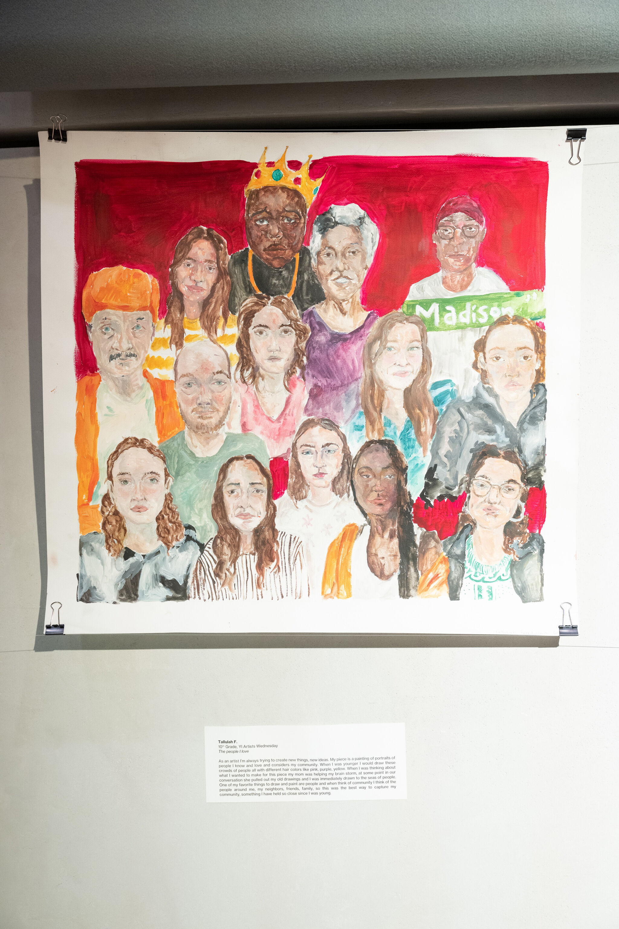 Colorful painting of diverse people, some wearing crowns and hats, displayed on a wall with a description below.