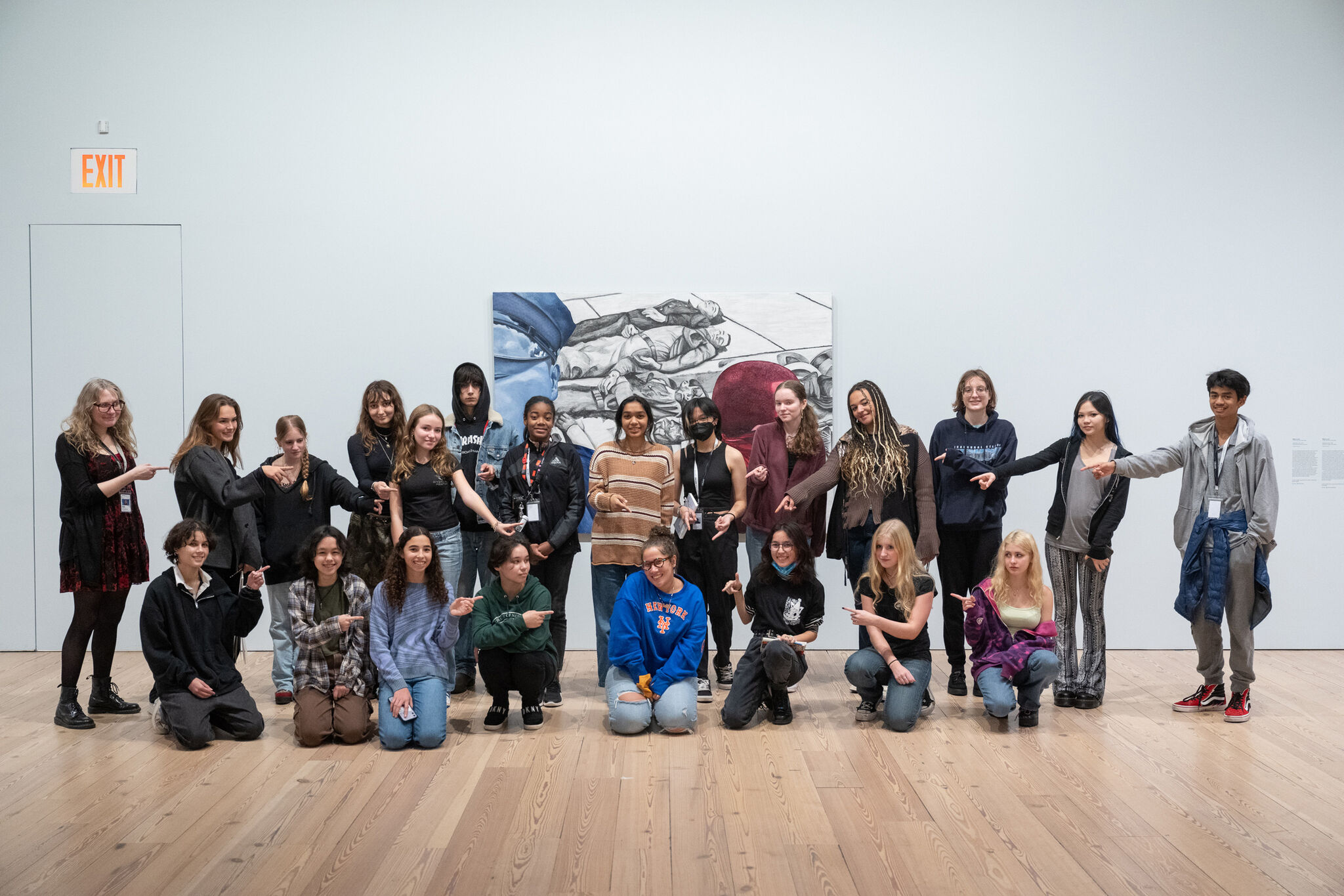 A group of people posing in front of a painting in a gallery, some kneeling, others standing, all pointing towards the center.