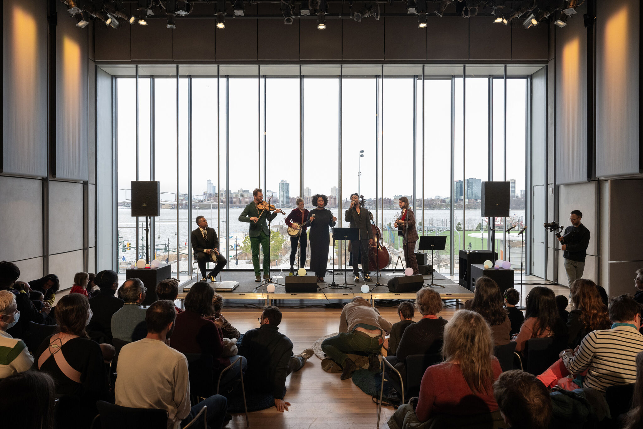 A jazz ensemble from Jazz at Lincoln Center is performing an all-ages concert at a Free Second Sunday event at the Whitney Museum. The group, consisting of musicians playing the violin, banjo, double bass, guitar, and percussion, along with a vocalist, is on a stage set against a large window overlooking the waterfront and city skyline. The audience, made up of a diverse group of attendees, is seated and attentively watching the performance, with some sitting on the floor. A videographer is capturing the event from the side. The space is illuminated by natural light, creating a warm and inviting atmosphere.