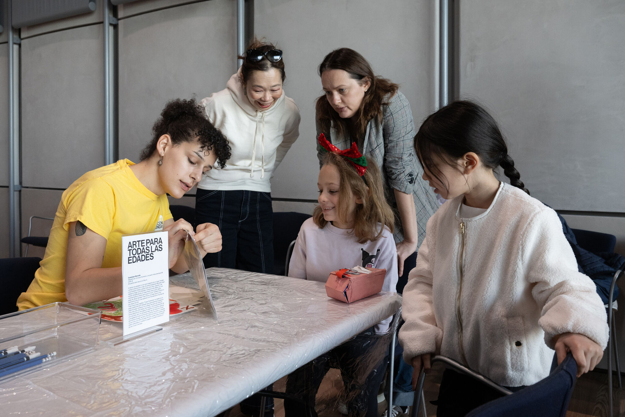 A facilitator in a yellow T-shirt leads a group in an artmaking program. Two children, one wearing a headband with festive decorations and another in a white jacket, look on while two older participants lean in, observing with interest. The setting is a well-lit indoor space with a table covered in plastic, art supplies, and an informational sign that reads “Arte Para Todas Las Edades” (Art for All Ages). The atmosphere is warm and collaborative, reflecting an intergenerational learning experience.