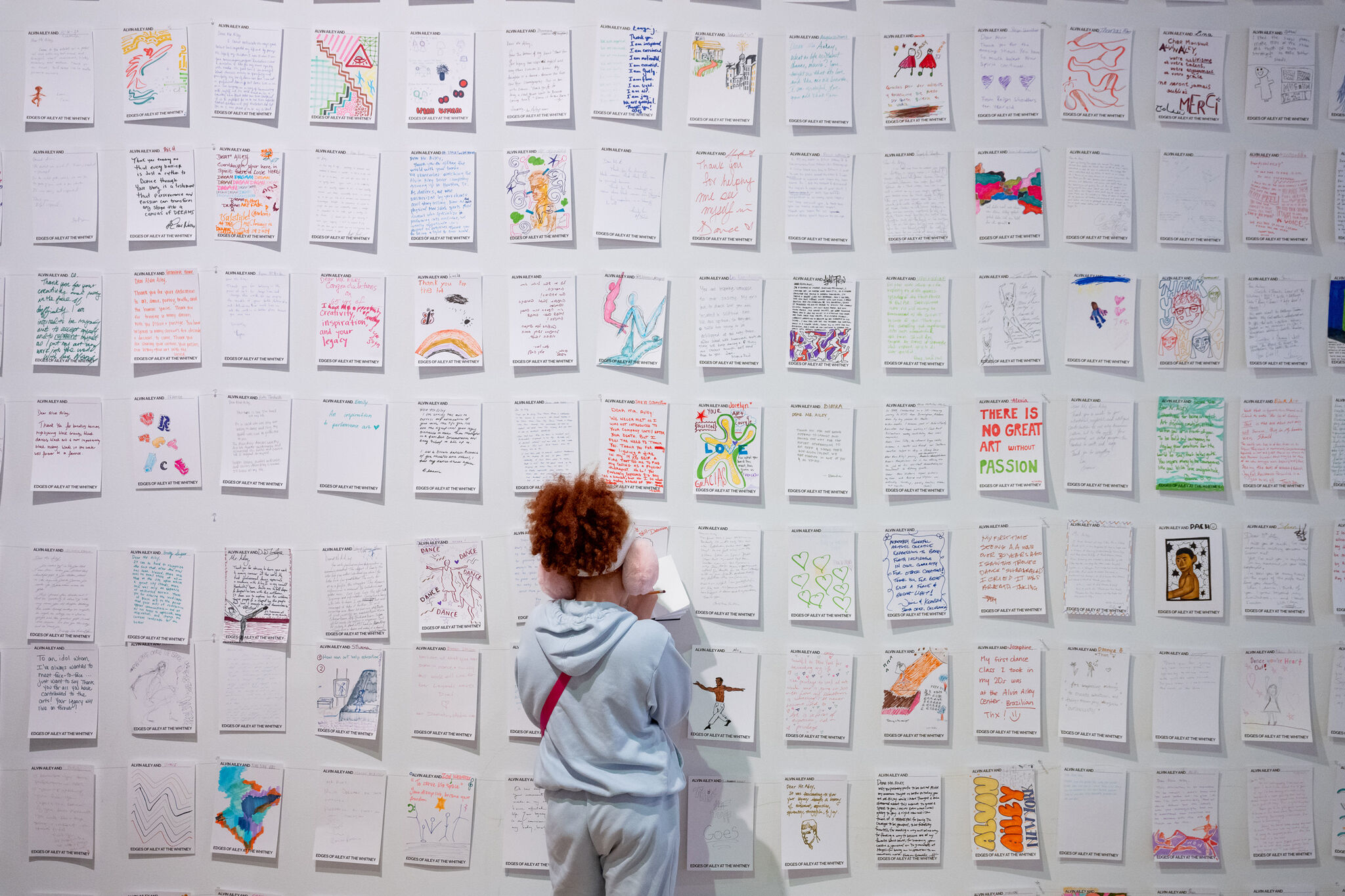 A white gallery wall is covered in a grid of handwritten and illustrated pieces of paper, each uniquely decorated with colorful drawings, text, and personal reflections. A viewer, dressed in a light-blue hoodie and wearing pink earmuffs, faces the wall, contributing to the display by adding another piece to the growing collection of community voices and artistic expressions. 