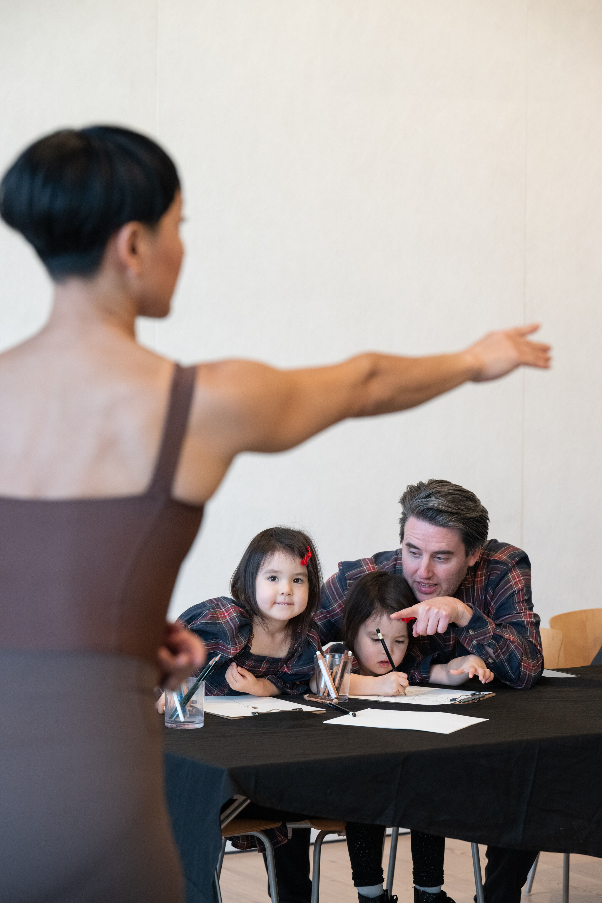 A facilitator in a yellow shirt is engaging a group in an art-making program at a Free Second Sunday event at the Whitney Museum. Two younger participants, one wearing a headband with festive decorations and another in a white jacket, are interacting with the activity, while two older participants lean in, observing with interest. The setting is a well-lit indoor space with a table covered in plastic, art supplies, and an informational sign that reads "Arte Para Todas Las Edades" (Art for All Ages). The atmosphere is warm and collaborative, reflecting an intergenerational learning experience.