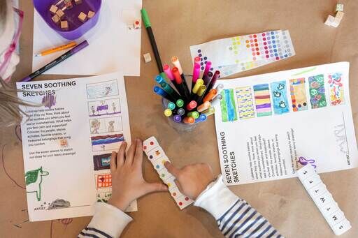 Child drawing on paper titled "Seven Soothing Sketches" with colorful markers and a pill organizer on a table.