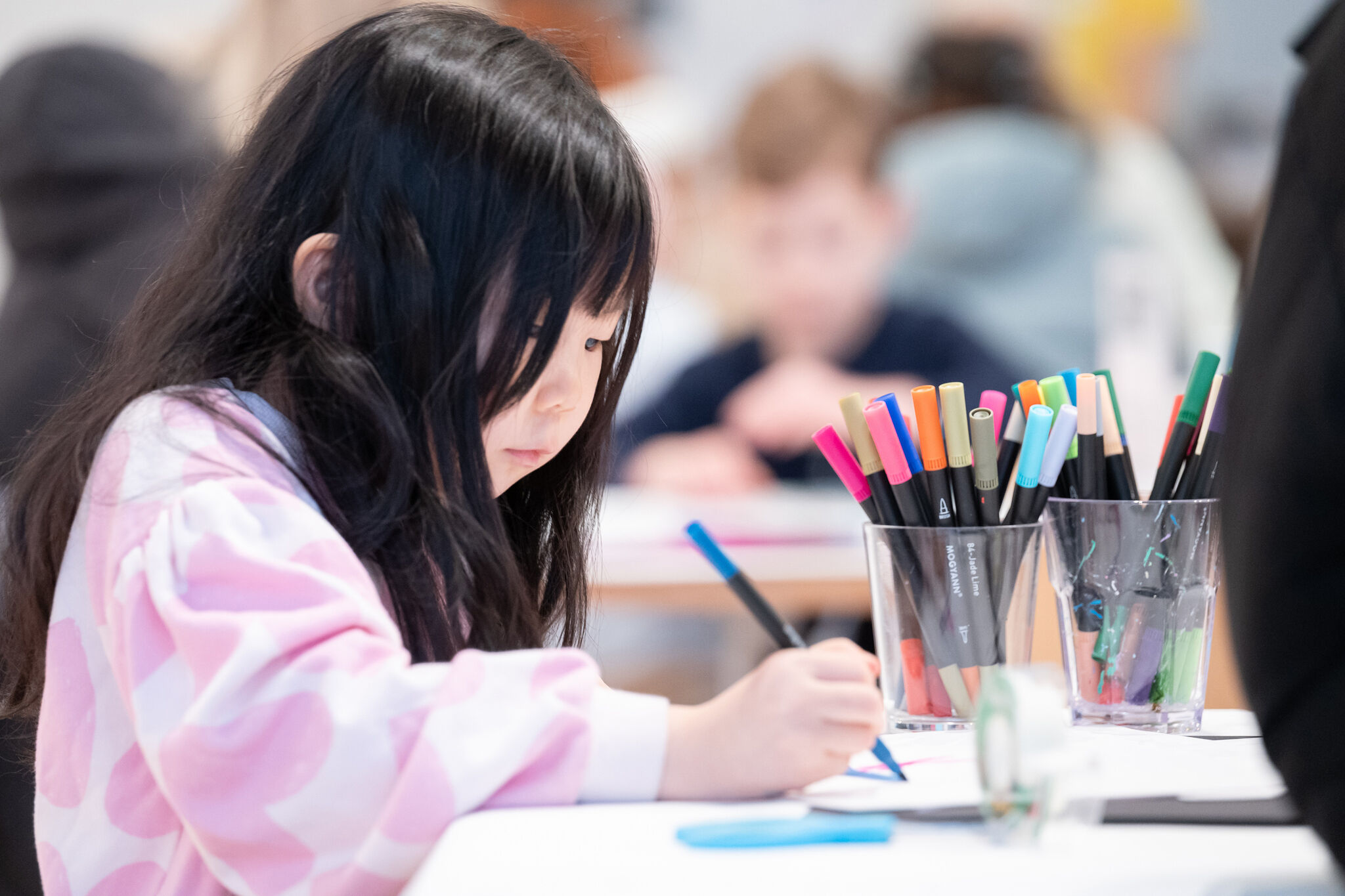 Young girl drawing with markers at a table, surrounded by colorful pens in cups.
