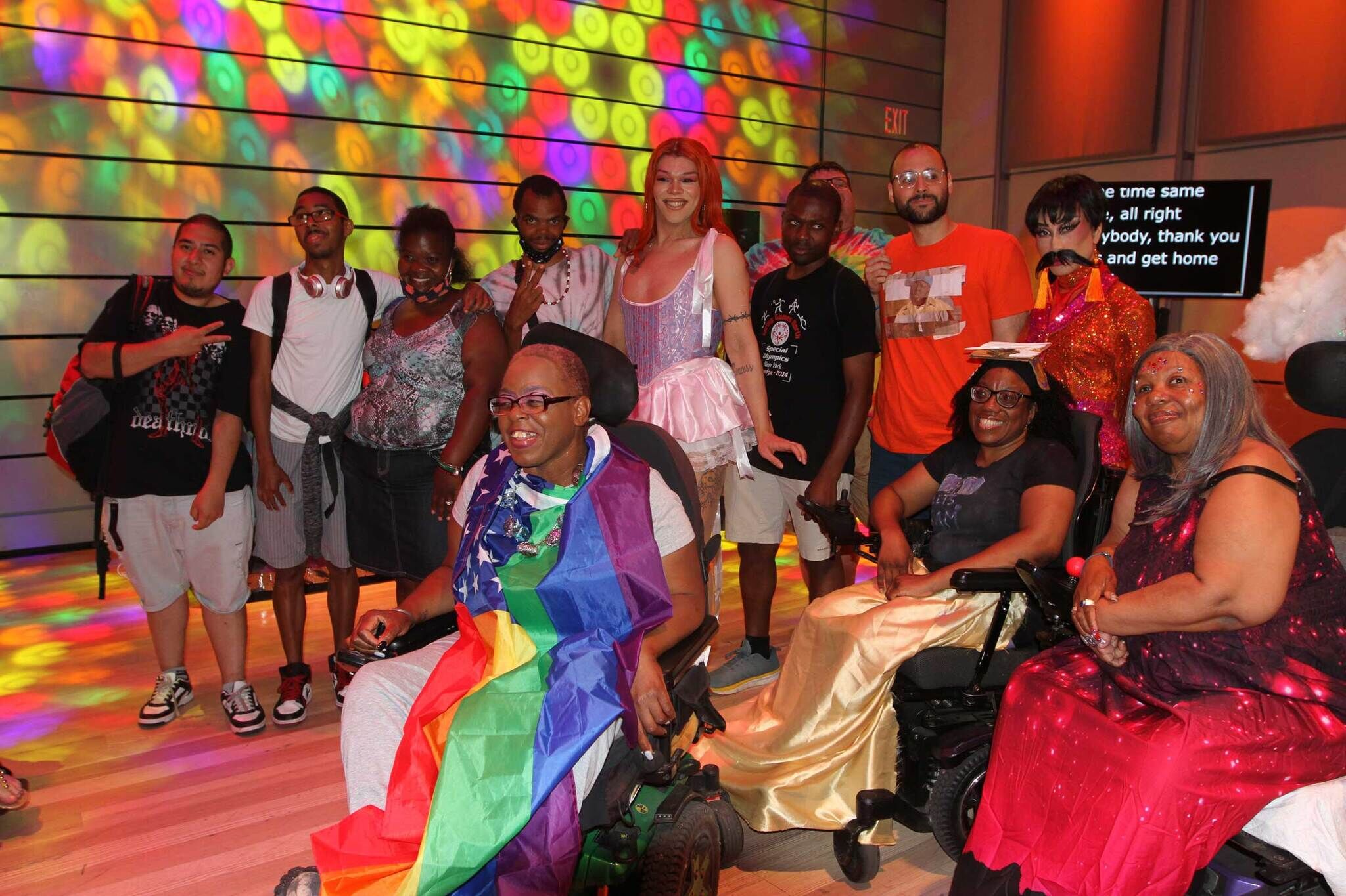 A group of people, some in wheelchairs and colorful outfits, smile together under bright, rainbow lights.