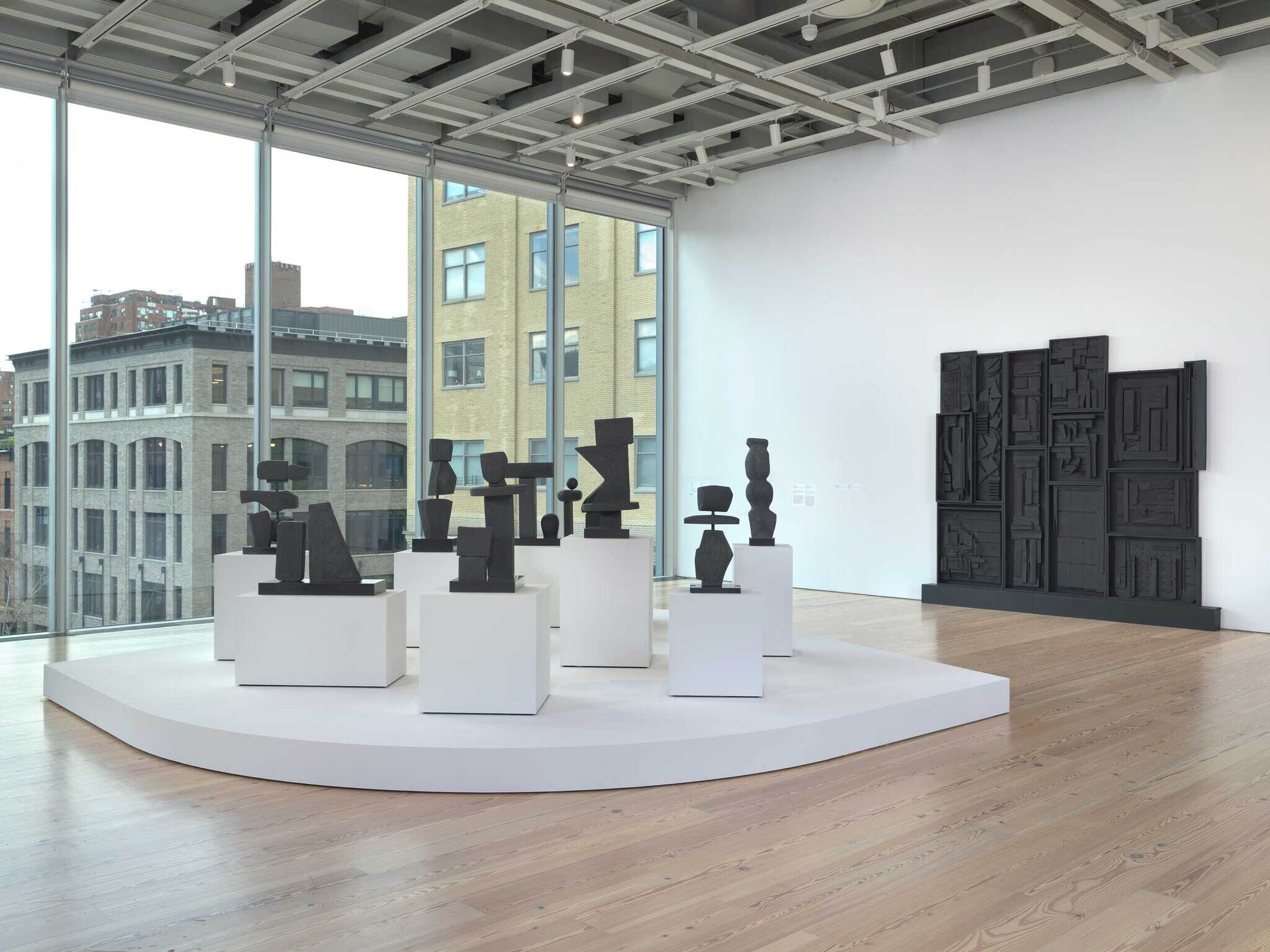 Several abstract black sculptures are displayed on white stands in a bright, modern art gallery with large windows.